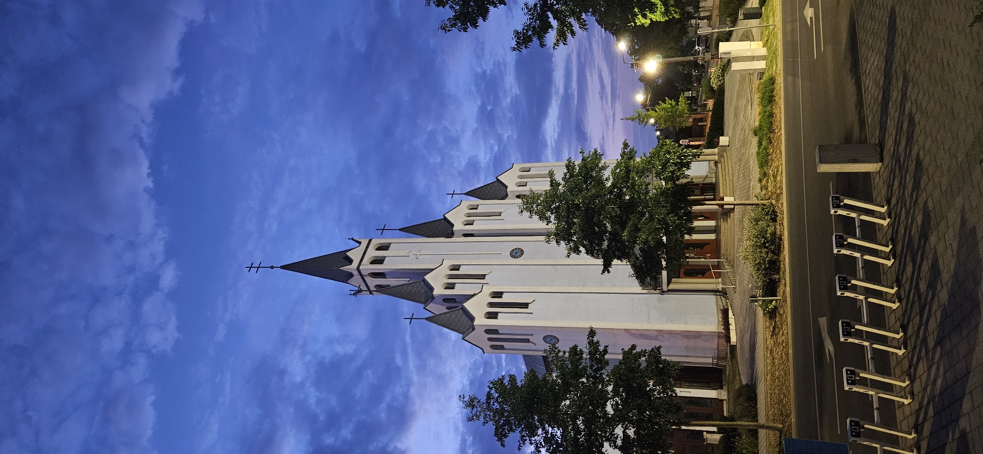 A white church with a tall spire and multiple smaller spires stands against a twilight sky with dramatic clouds. The church features arched windows and crosses atop its spires. In front of the church, there are trees and a paved walkway leading to the entrance. A street runs in front of the church, with streetlights illuminating the scene. A bike rack is visible on the sidewalk. The image was taken in the city of Hévíz, Hungary.