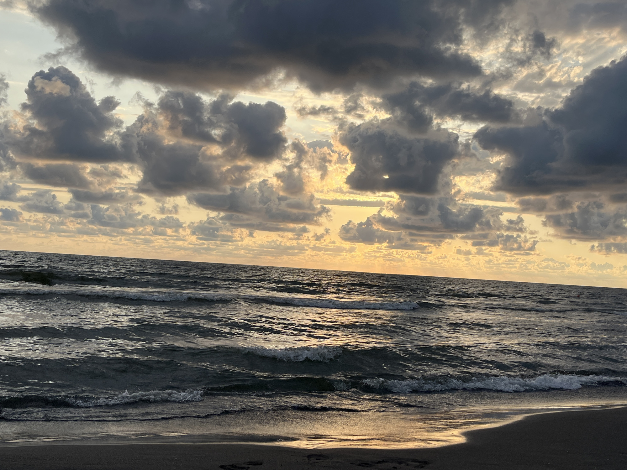 The image depicts a beach scene at sunset. The sky is filled with dramatic, dark clouds interspersed with patches of golden light, suggesting the sun is either rising or setting. The sea is choppy with visible waves breaking on the shore, and the wet sand reflects the warm sky colors. There are no people or discernible objects in the scene besides the natural elements of sky, sea, and sand. The location is identified as a beach in the village of Supsa, Georgia, part of the Supsa community.
