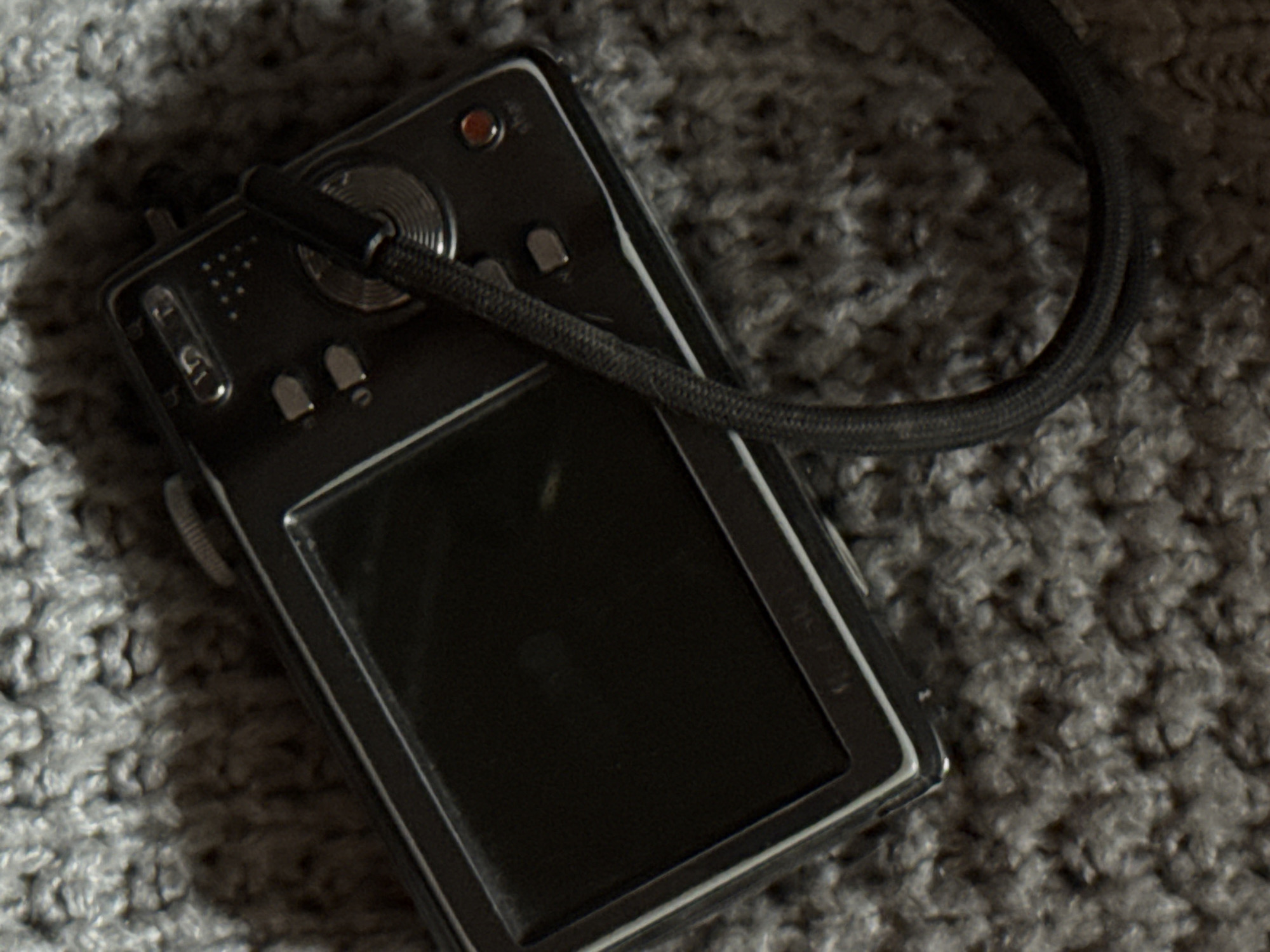 The image shows a black digital camera lying on a textured gray surface, possibly a knitted fabric or carpet. A black strap is attached to the camera and extends across the frame. The camera has several buttons and a screen visible. A red circular button is located on the top left of the camera. To its right, there is a silver dial. Below the dial, there are several small buttons arranged in a cluster, and a larger rectangular button. The camera has a prominent black screen in the center. The overall lighting suggests an indoor setting, likely a dimly lit room, as there are shadows cast on the surface. No specific time of day, weather conditions, or location cues other than the provided context of Izhevsk, Russia are discernible from the image. No legible text is visible.