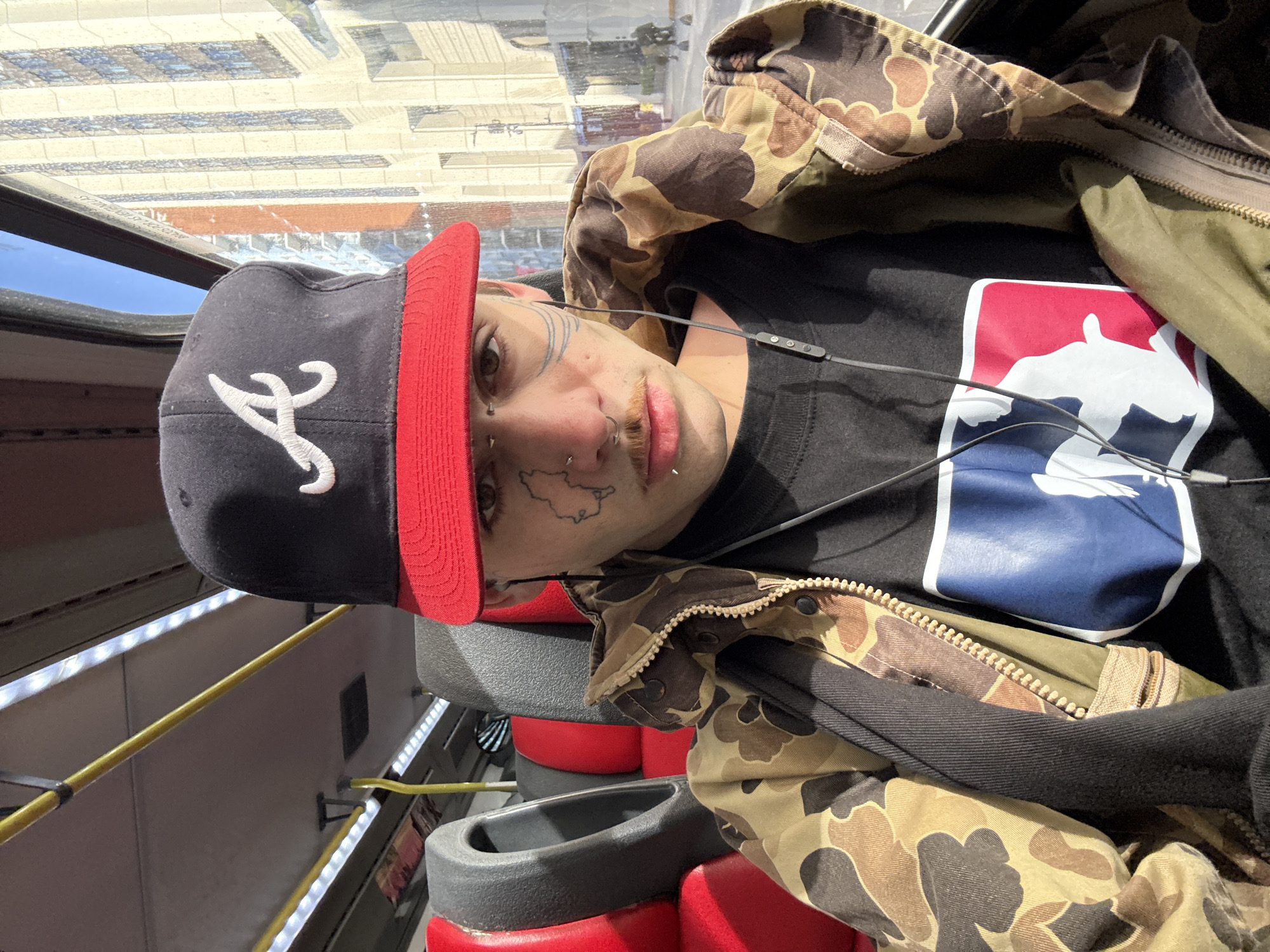 A young person is captured in a selfie, looking directly at the camera while seated inside what appears to be a public transport or tour bus. They are wearing a black baseball cap with a red brim and a white stylized 'A' logo, a black t-shirt featuring the Major League Baseball (MLB) logo, and a camouflage-patterned jacket. Earbuds with an inline control panel are draped around their neck.
The person exhibits multiple facial piercings, including nostril rings, a septum piercing, and lip rings. Intricate linear tattoos adorn their face around the eyes, and a distinct map-like outline is visible on their cheek. They have light brown eyes and visible stubble.
The interior of the vehicle features red and black seats, a yellow grab bar, and illuminated LED strip lights along the side panel. Outside the window, parts of buildings with a grid-like pattern are visible against a clear blue sky, indicating daytime. The person maintains a neutral expression. Despite the presence of American sports logos on their clothing, the scene is situated in Buenos Aires, Argentina.