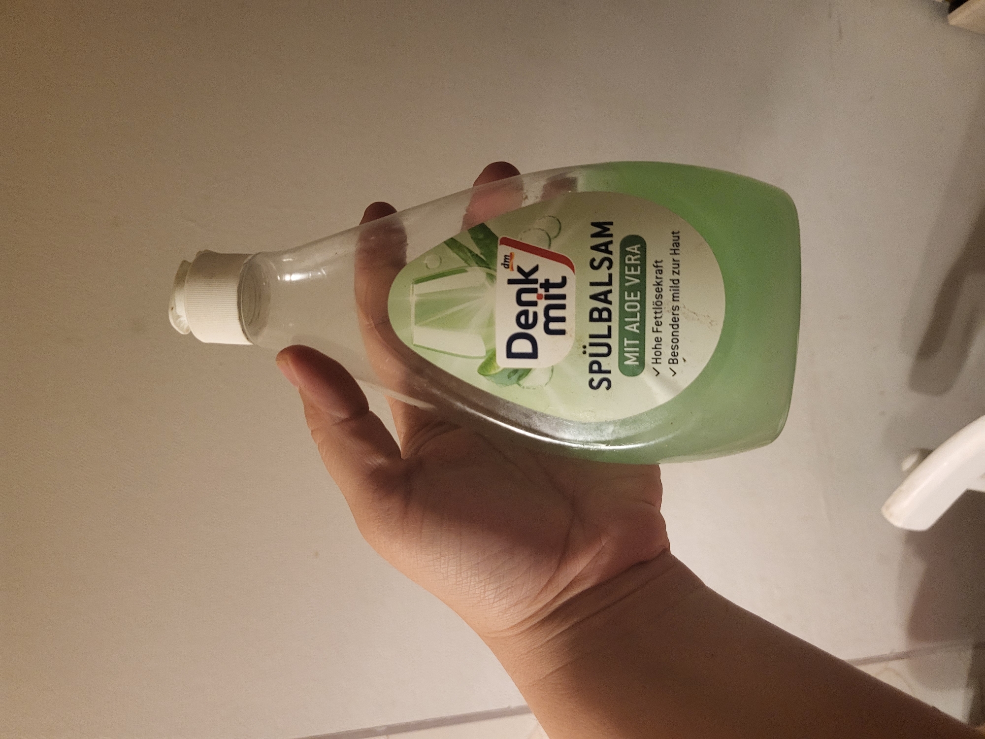 A person's hand is holding a clear plastic bottle of green dish soap. The label on the bottle reads "Denk mit SPÜLBALSAM MIT ALOE VERA" and features an image of aloe vera leaves. The background is a plain, light-colored wall. The image was taken in Pinneberg, Germany.