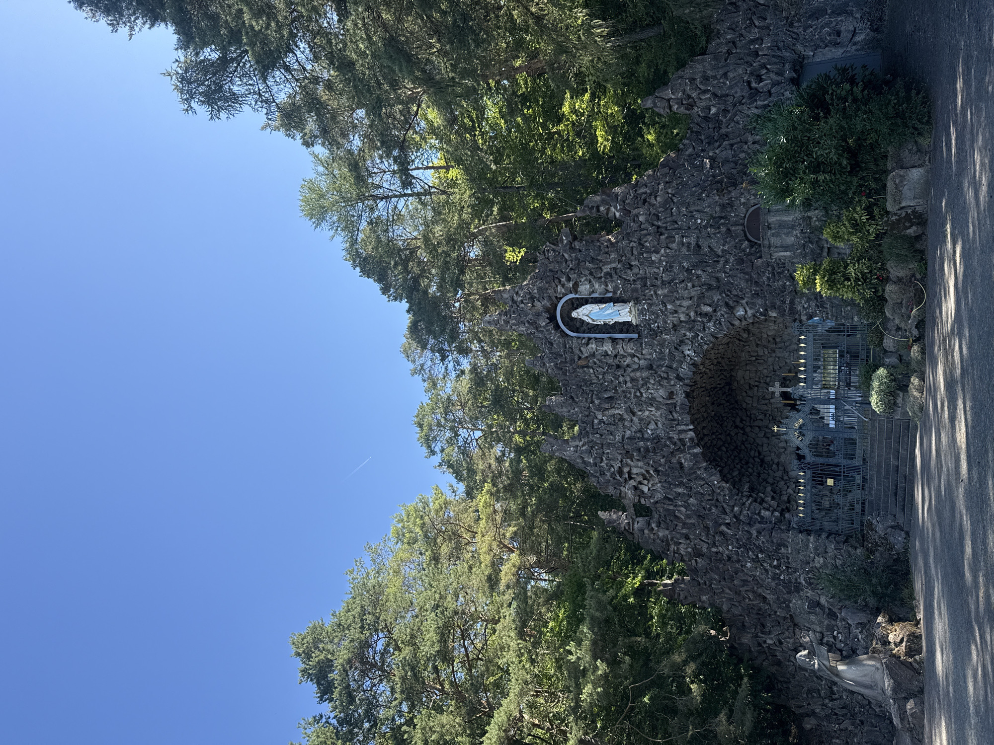 The image depicts a grotto-like structure made of dark, irregular stones, situated in a forested area with tall evergreen trees dominating the background against a clear blue sky. At the apex of the stone structure, a statue of the Virgin Mary, dressed in blue and white, is visible within a niche. Below this, a large arched opening leads to an area enclosed by ornate metal gates, behind which can be seen steps and what appears to be an altar with crosses. To the left of the entrance, a stone statue of a cloaked figure is positioned. The ground in front of the grotto is a dark, shaded pathway, with some vegetation and rocks around the base of the structure. This scene is identified as the Mariengrotte (Mary's Grotto) in Bad Salzschlirf, Germany.