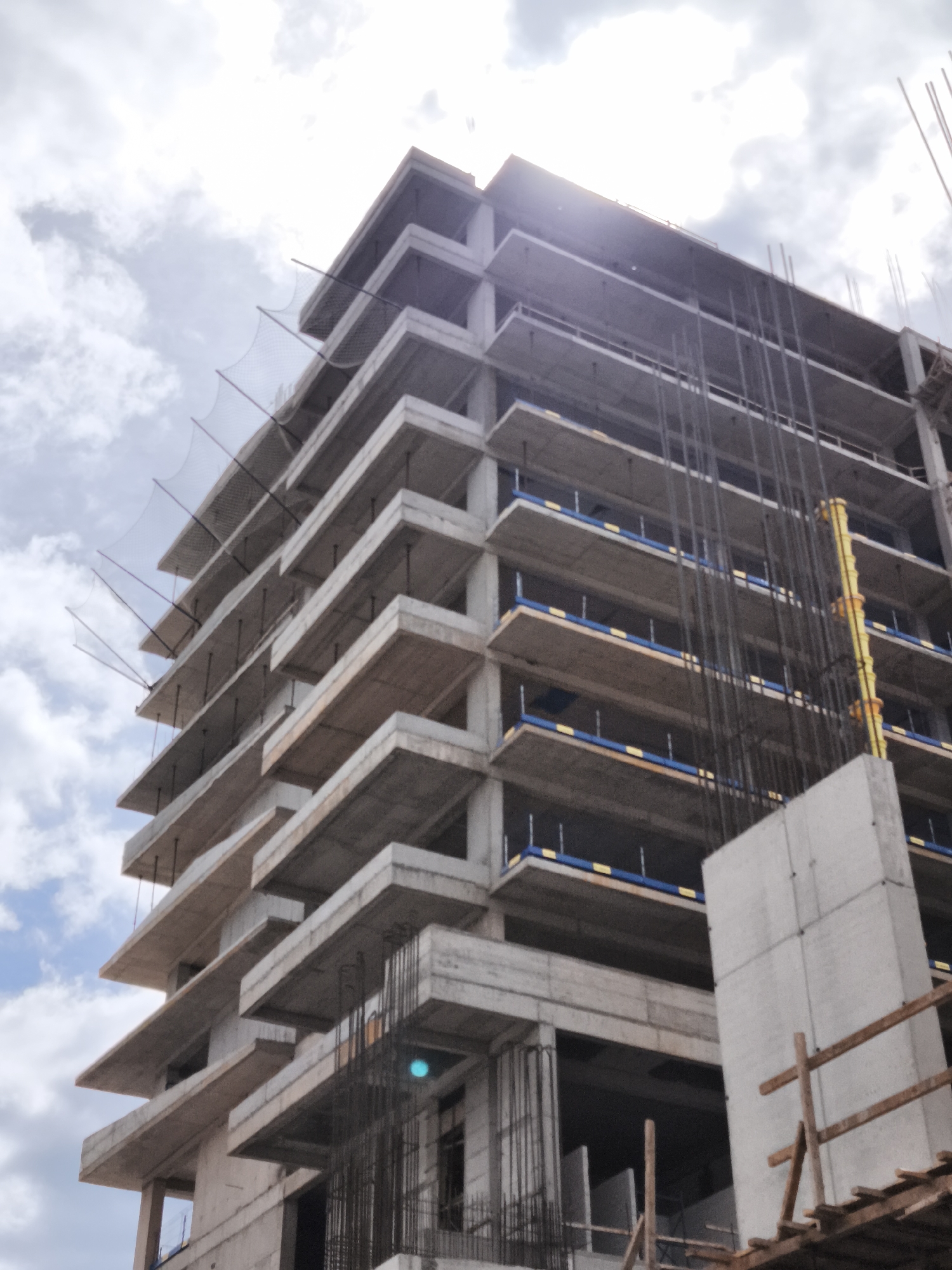 The image depicts an unfinished multi-story concrete building under construction. Located in Budva, Montenegro, the building features protruding concrete floor slabs and exposed rebar structures. Safety netting is visible on the upper floors. Construction materials, including scaffolding and yellow conduits, are present. The sky is cloudy, with the sun shining through. There are no people visible in the frame.