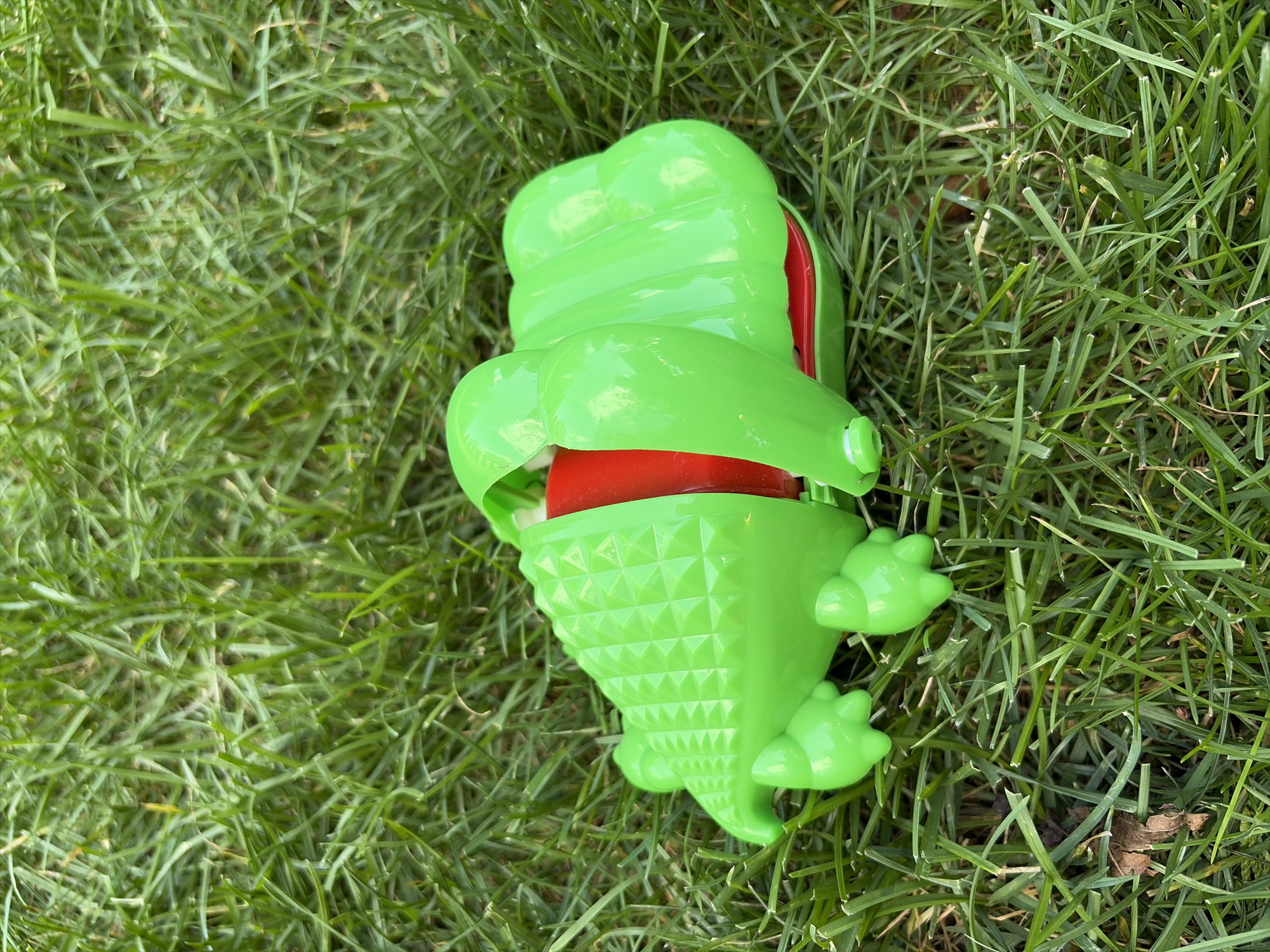A bright green plastic toy alligator lies on a patch of green grass. The alligator toy is split open, revealing a red interior. Its body is segmented and textured to resemble scales, and it has four stubby legs with three toes each. The toy is positioned horizontally on the grass. The background consists of the blades of green grass.
