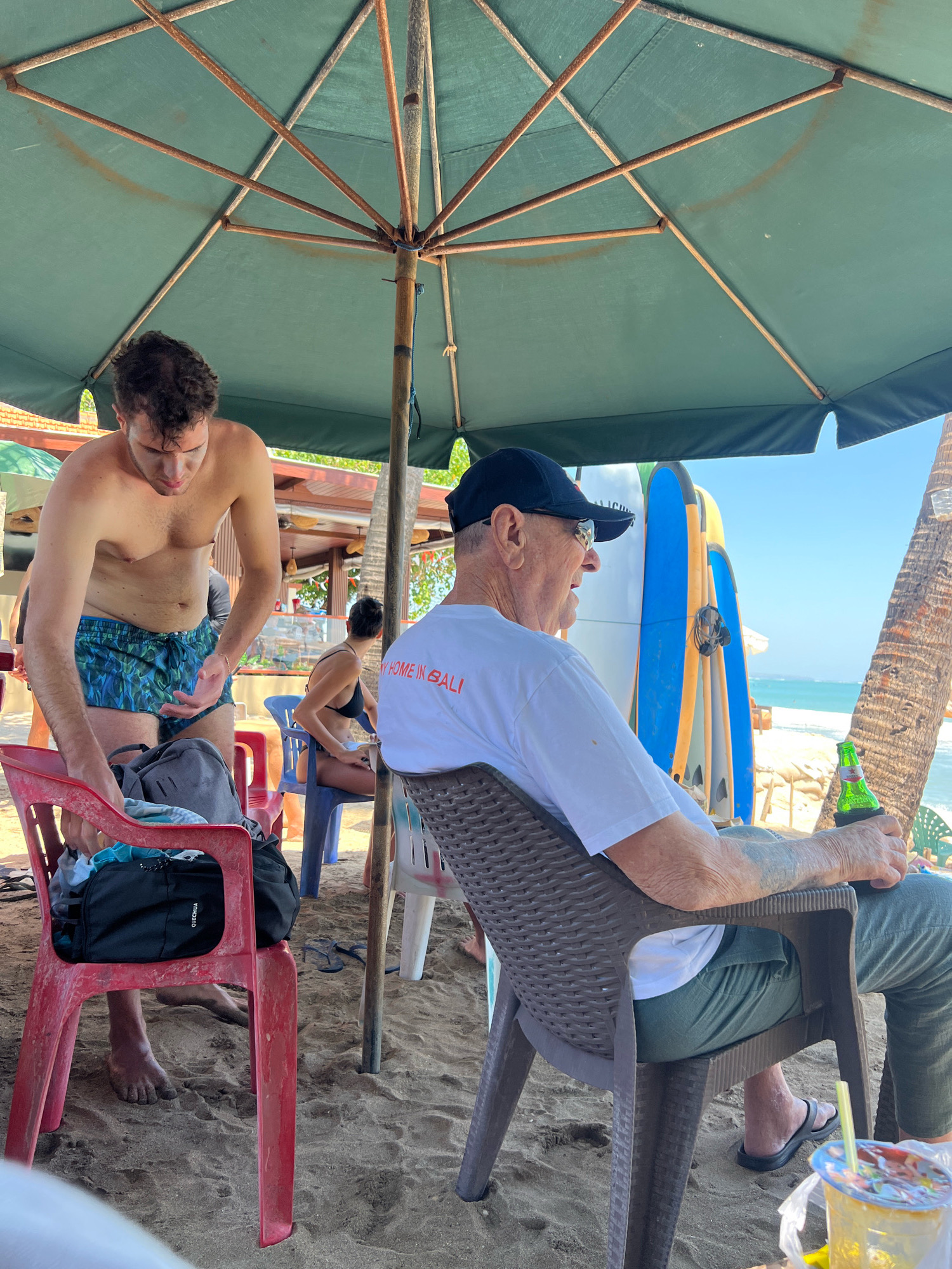 In Kuta, Indonesia, on a sandy beach under a large green umbrella, two men are seated in chairs. To the left, a younger man, shirtless and wearing patterned swim trunks, is reaching towards a backpack on a red plastic chair. To the right, an older man wearing a white t-shirt, dark baseball cap, and green pants is holding a green bottle with a koozie. The t-shirt has text that reads "MY HOME IN BALI". In the background, surfboards are propped upright near the water's edge. The environment is sunny with a clear blue sky.