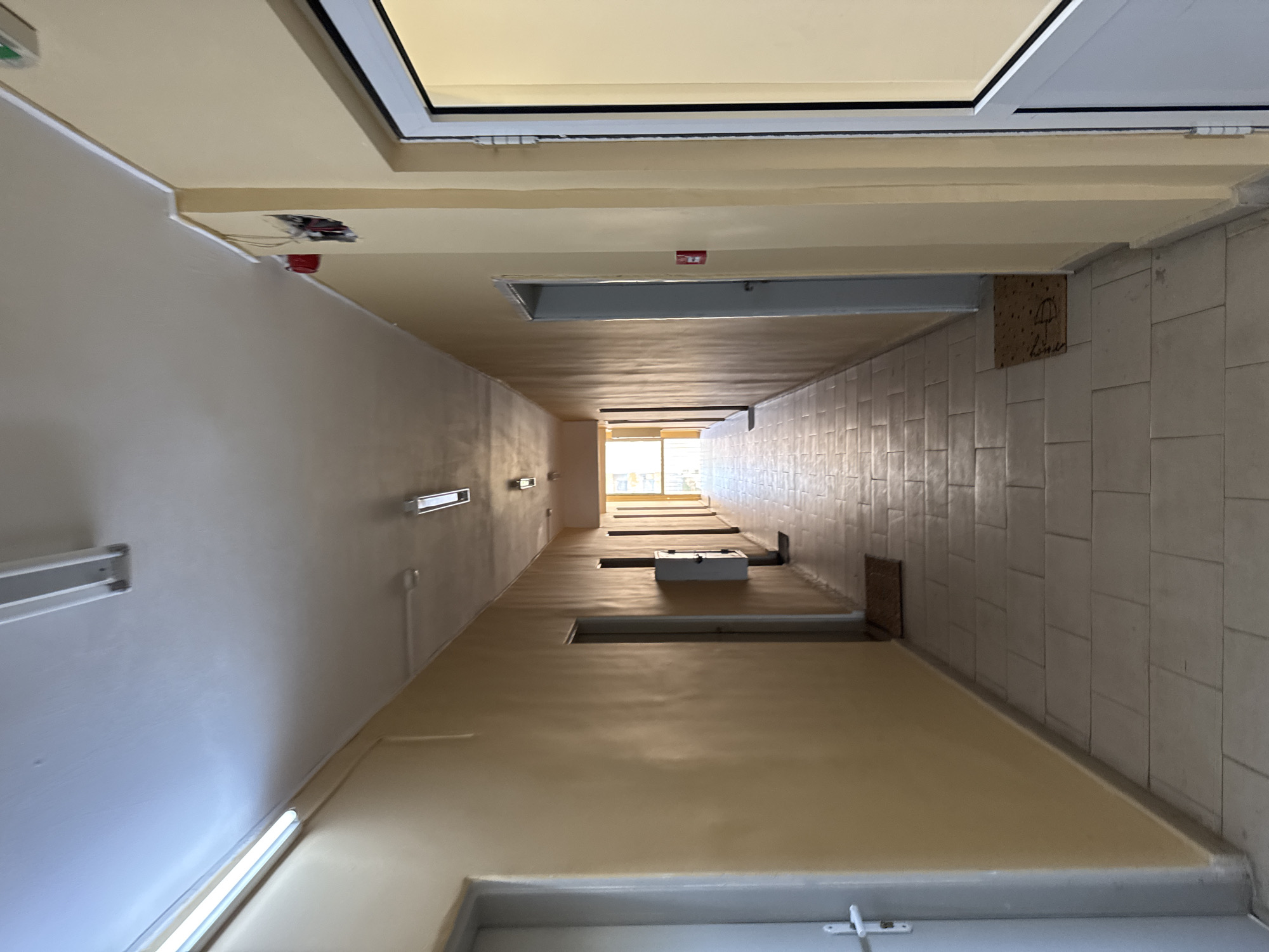 The image shows a long, narrow corridor with cream-colored walls and a tiled floor. Fluorescent lights are mounted on the ceiling. Several doors are visible along the walls. At the far end of the corridor, natural light streams in from an opening. There are no people or animals visible in the image. This scene is consistent with a hallway in a residential building or an institutional setting. Without further information, it is impossible to determine the specific location in Sofia, Bulgaria.