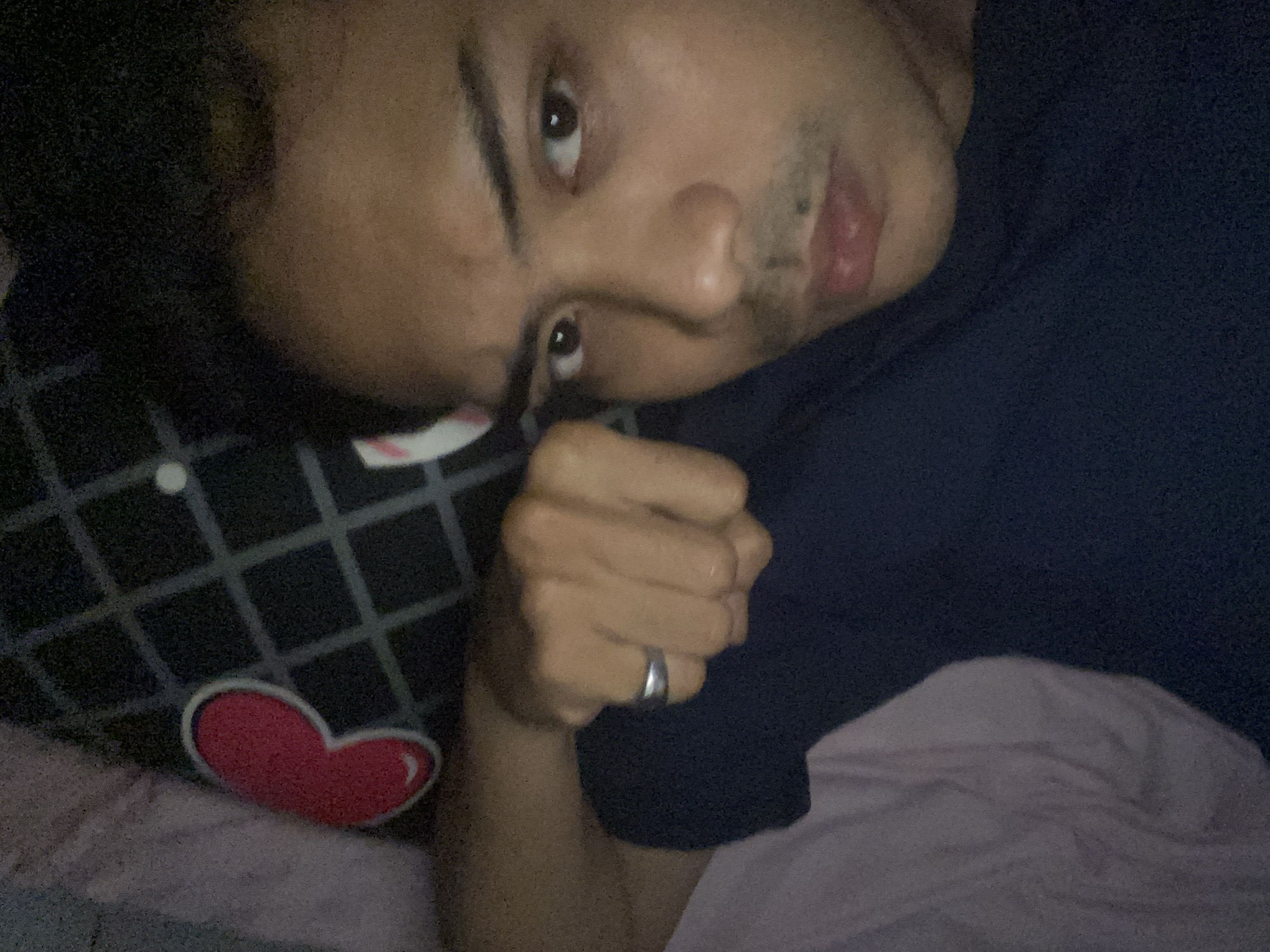 The image shows a close-up selfie of a man lying down, possibly in bed. He is wearing a dark blue t-shirt and has a silver ring on his left hand, which is held up towards his face, partially obscuring his chin. The background includes a pillow with a red heart design and a grid pattern, and a crumpled light-colored fabric, likely bedding. The man's expression is neutral, with his eyes looking directly at the camera. The lighting is dim, suggesting it might be nighttime or an indoor setting with low light. There is no discernible information that would indicate the location within Dukuh, Indonesia.