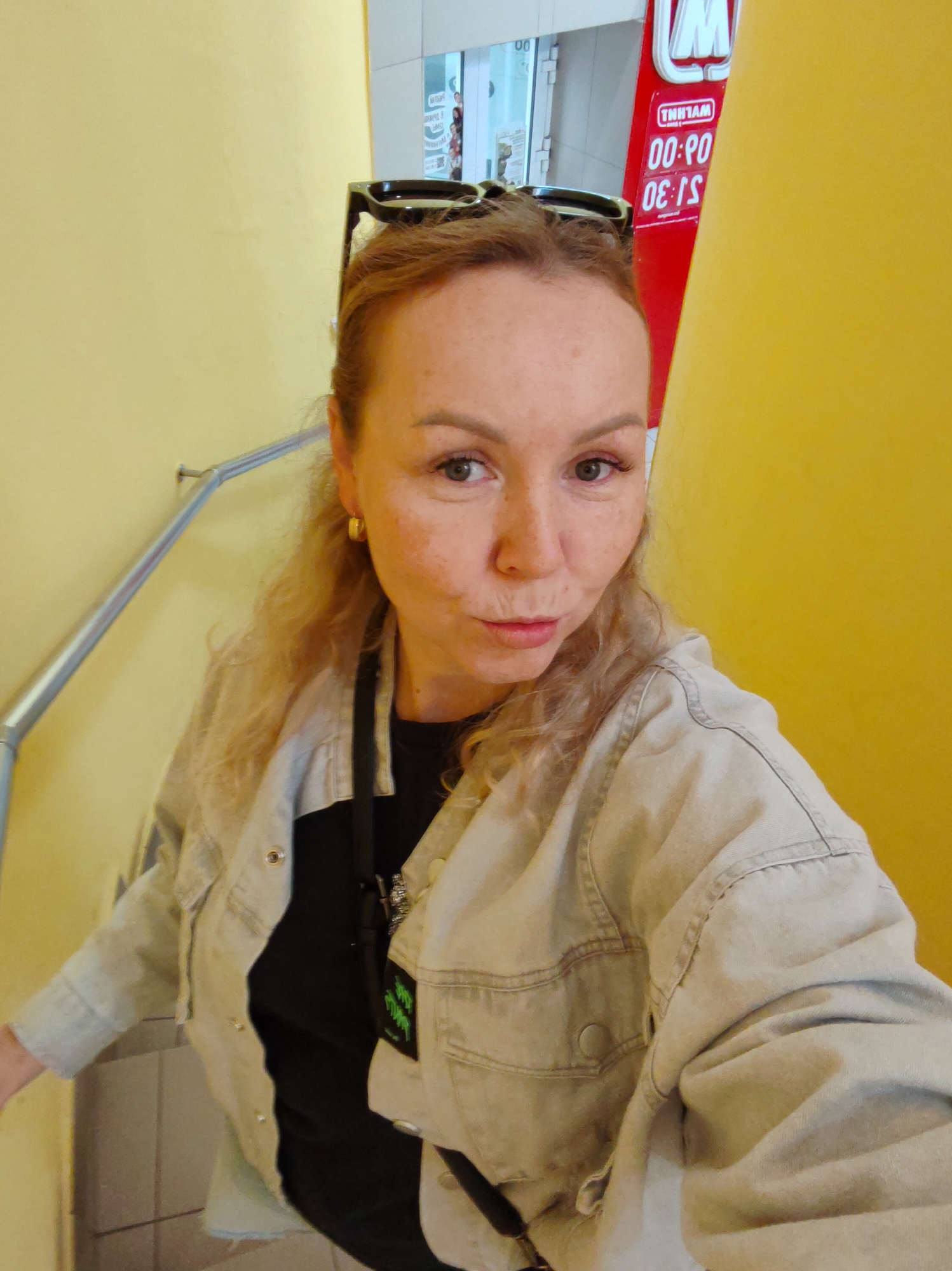 A blonde woman with freckles, wearing a light-colored denim jacket over a black t-shirt and gold hoop earrings, is taking a selfie. Sunglasses are perched on top of her head. She is standing on a staircase with a yellow wall on the right and a metal railing on the left. In the background, a red sign with white text and a logo is visible. The text on the sign appears to be in Russian and is reversed, indicating it's a reflection or a sign viewed from the other side. The environment suggests an indoor public space, possibly a commercial area. The timestamp "00:00" and "06:30" are legible on the sign.