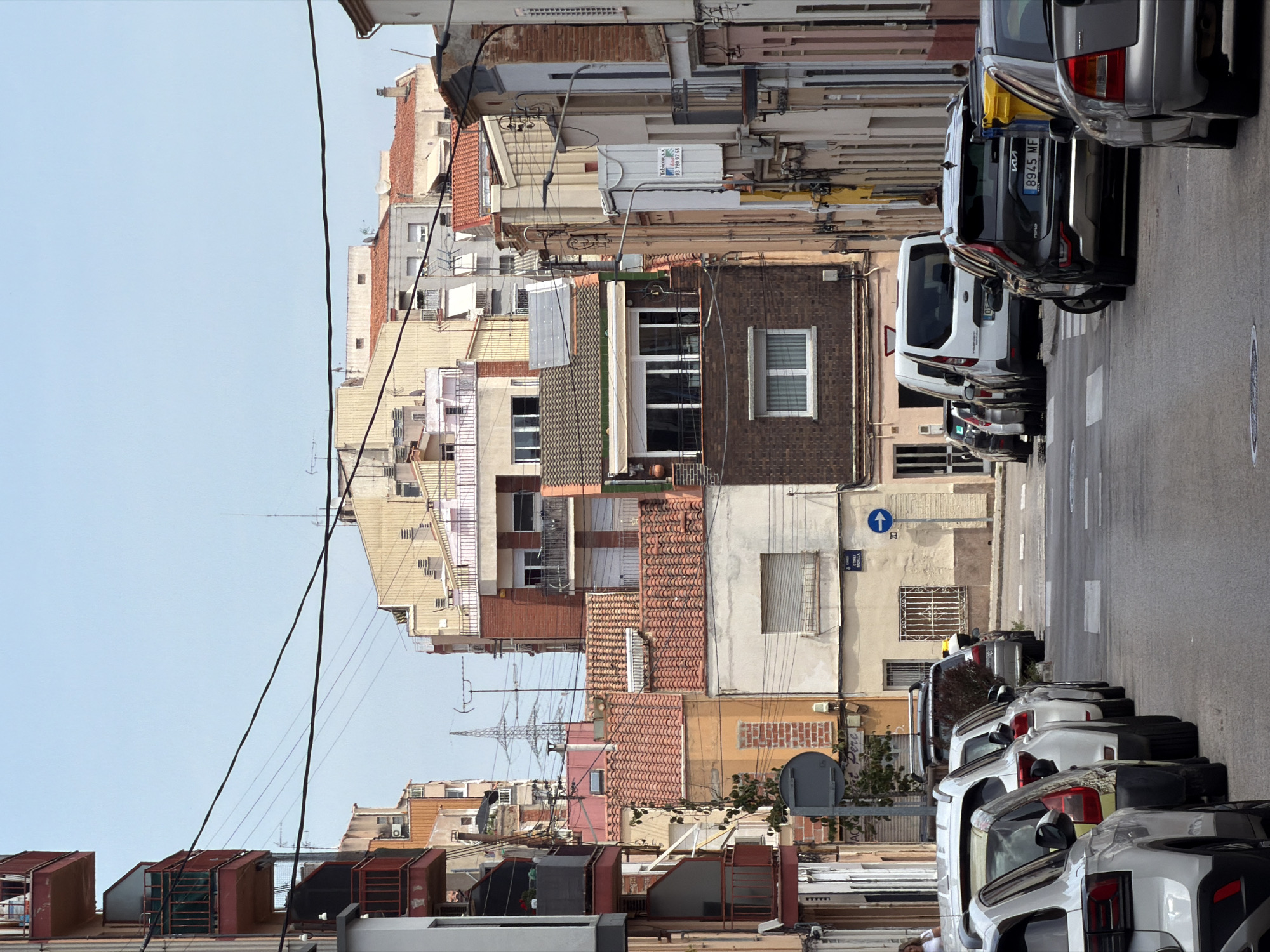 The image shows a street scene in Terrassa, Spain, with buildings lining both sides of the road. The architecture appears to be a mix of residential and commercial buildings, with various styles and colors. Several cars are parked along the street, and some people are visible walking or standing. The sky is clear, suggesting a sunny day. The scene depicts a typical urban environment with everyday activities taking place.