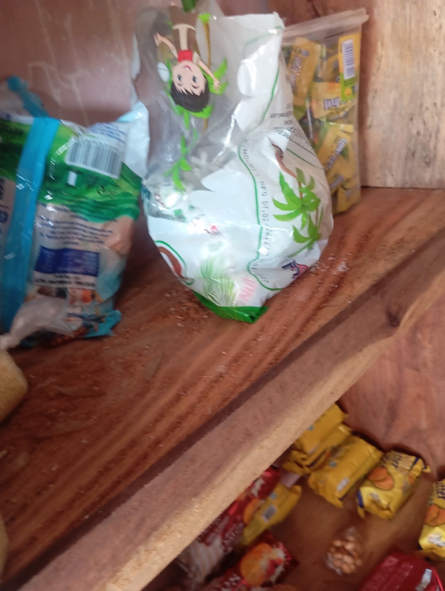 The image depicts a shelf inside a room, likely a home or small shop, in Jalingo, Nigeria. The shelf is made of wood and appears somewhat worn. On the shelf are several food items, primarily packaged snacks. To the left, a large, partially opened bag of what appears to be rice or flour is visible, with blue and green accents. In the center, a clear plastic bag is filled with small, individually wrapped yellow candies. To the right, a collection of individually wrapped snacks, mostly yellow and red, are arranged. Some of these appear to be biscuits or crackers. A few loose nuts or seeds are also scattered on the shelf. The lighting is dim, suggesting an indoor environment. The overall scene is a still life arrangement of food products on a wooden surface.