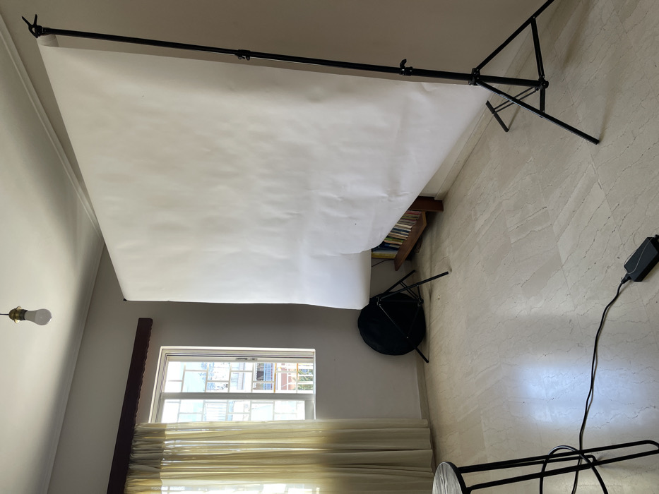 The image displays an indoor room, oriented as if rotated 90 degrees clockwise, showing a photography backdrop setup. When viewed upright (rotated 90 degrees counter-clockwise), the scene reveals a light-colored room with a white paper or fabric photography backdrop suspended from a black metal support system.
The backdrop system consists of a horizontal black bar held up by two vertical stands, one visible on the far left and the other on the far right. The white backdrop material hangs from this bar, cascading downwards and extending onto the floor, creating a seamless background. The backdrop appears somewhat wrinkled and has a few minor blemishes.
On the left side of the room, a window with multiple panes is visible towards the bottom. Light-colored, possibly sheer, curtains cover the lower half of the window, allowing daylight to filter through. Outside the window, glimpses of other buildings can be seen, indicating an urban or residential setting. Above and to the left of the window, a bare white light bulb in a simple, dark fixture hangs from the ceiling.
Towards the right side of the room, under the draped backdrop, a wooden bookshelf filled with books is partially visible. In front of the bookshelf, a small, dark object that resembles a collapsed tripod or a small, dark stool is present.
The floor of the room is covered with light-colored tiles, possibly marble or a similar material, exhibiting subtle patterns or veining. In the bottom right corner of the image, a black power adapter with a black cable extending from it is visible on the tiled floor. Part of a black metal leg, likely belonging to another stand or piece of photography equipment, is also seen in this area.
The walls are painted a light, neutral color. The overall impression is that of a functional space temporarily set up for photography, combining elements of a home environment with specialized equipment. The time of day appears to be daytime, judging by the light coming through the window.
