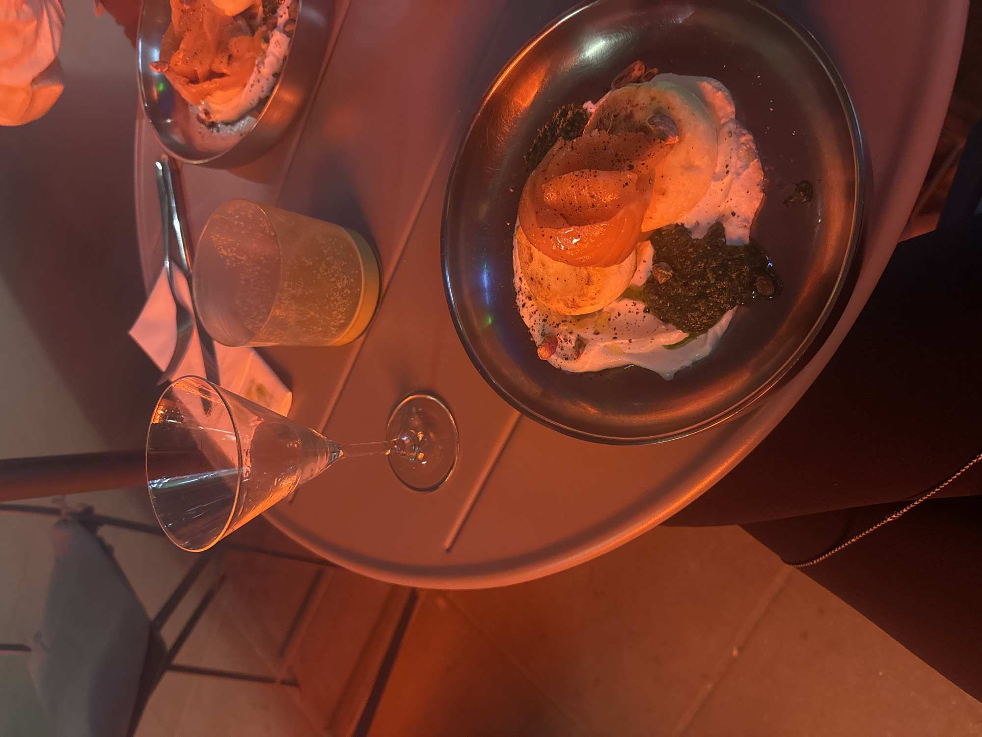 A high-angle shot shows a gray, round outdoor table with two metal plates, a tall cylindrical glass of yellow liquid, and an empty wine glass on it. The plate on the right contains food that appears to be layered, consisting of a white creamy base, a dark green substance, and orange slices on top, sprinkled with black pepper. The plate on the left has a similar dish. A napkin and silverware are also present. The table is situated outdoors, likely at a restaurant or bar, indicated by the chair leg visible at the bottom left. A person dressed in black clothing is partially visible on the right side of the frame. The lighting is warm, possibly from an overhead light or sunset. While the exact location in Warsaw, Poland, cannot be determined from the image alone, it depicts a dining experience in an outdoor setting.