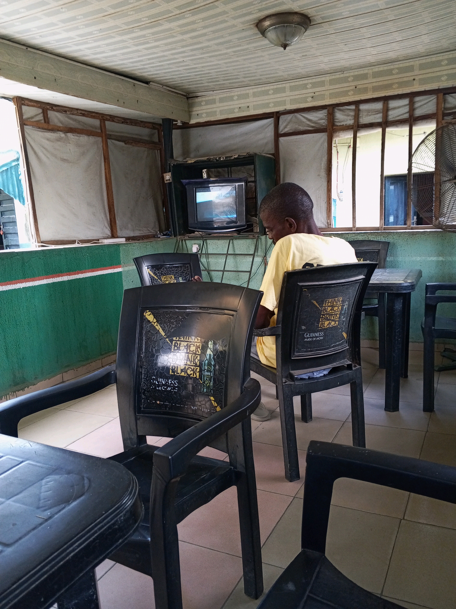 The image depicts an interior scene, possibly a bar or restaurant, in Zango-ndaji, Nigeria. A man wearing a yellow t-shirt is seated in a black plastic chair, facing away from the camera and towards a television screen. The television is a CRT model, displaying an image. The man appears to be observing the screen. Several other black plastic chairs and tables are visible in the room, some with a "Guinness" branded advertisement on the backrests. The walls are painted green with a red and white stripe. The ceiling is made of white panels. Natural light enters from windows covered with light-colored fabric. A large industrial fan is visible in the background. The flooring is tiled.