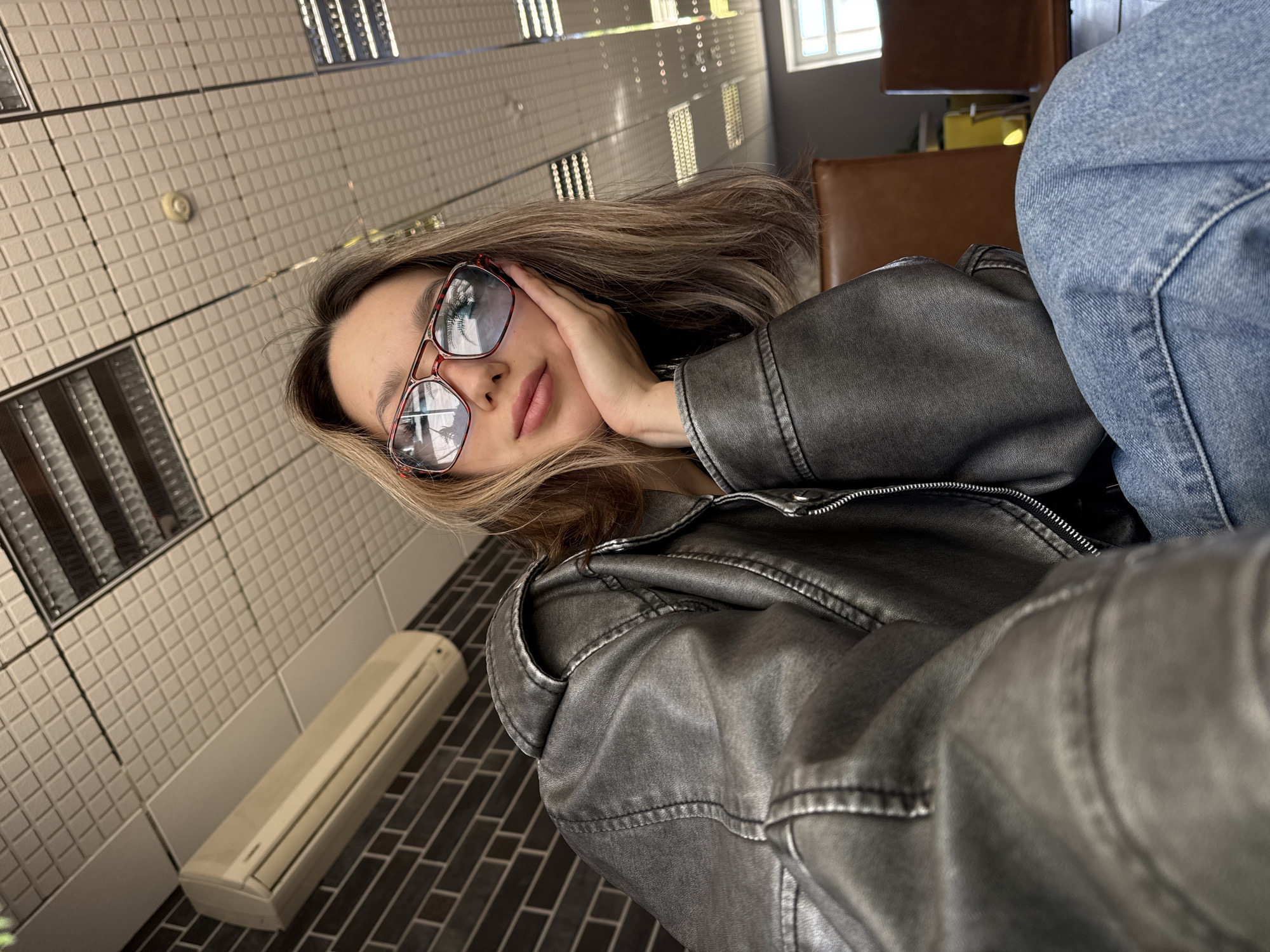 A woman with blonde hair and red glasses is sitting in an unidentified location in the city of Unknown, Unknown. She is wearing a grey leather jacket and blue jeans. She is resting her face on her hand. The background features white tiled walls with narrow rectangular mirrors and a white air conditioning unit. The floor is tiled in dark brown. Brown chairs can be seen in the background, indicating a cafe or restaurant setting. The overall lighting suggests an indoor environment.