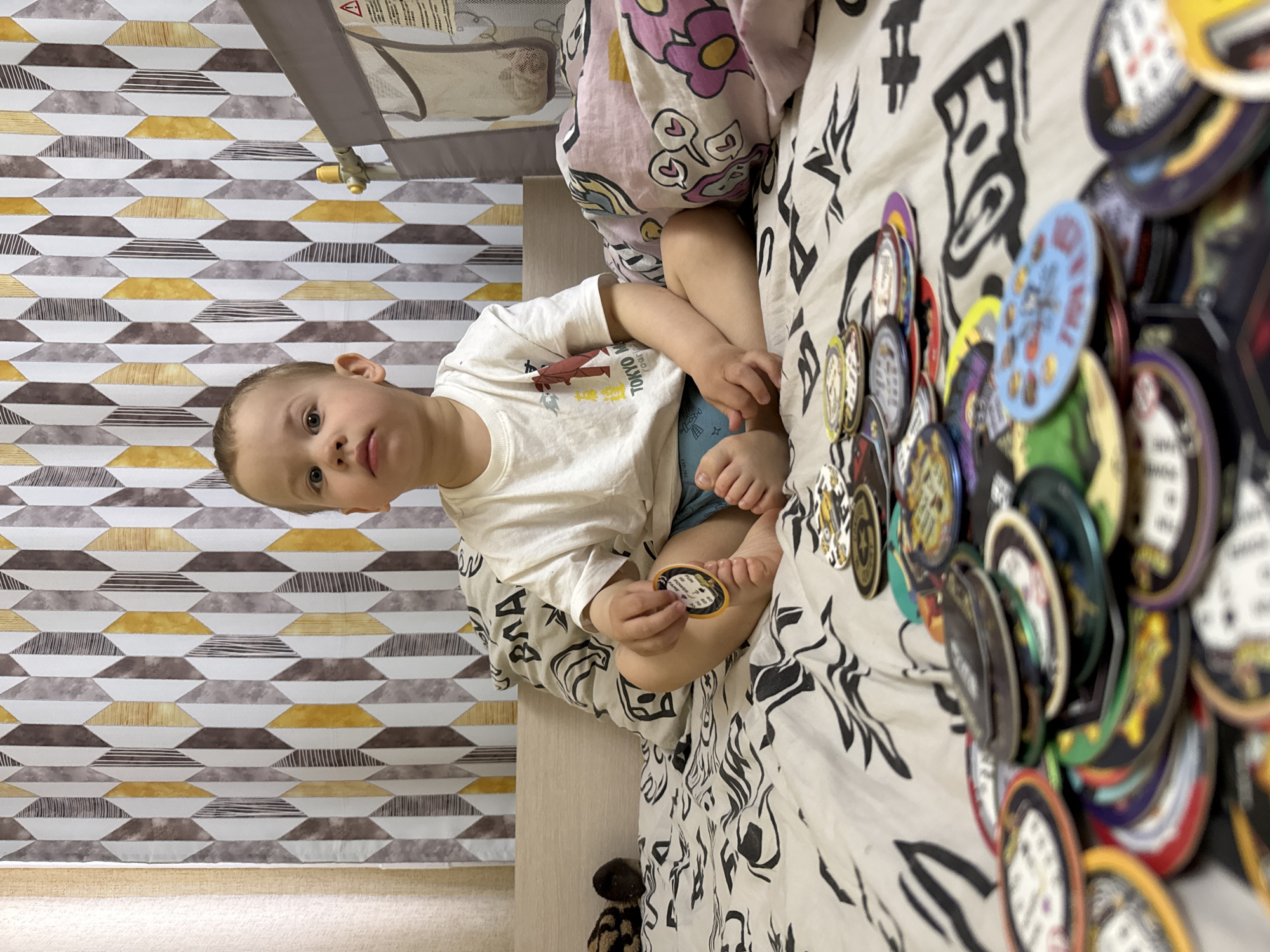 A young child is sitting on a bed with a pile of round collectibles, possibly POGs or similar tokens. The child wears a white t-shirt and blue shorts, and holds a round token in their hand. The bed has a patterned black and white sheet, and there is a blanket with hearts on it. Next to the bed is a crib with a patterned backdrop in white, gray, brown, and yellow geometric shapes. The location is not discernible from the image, so the presence of a specific city in Russia like Rusanovo cannot be verified.