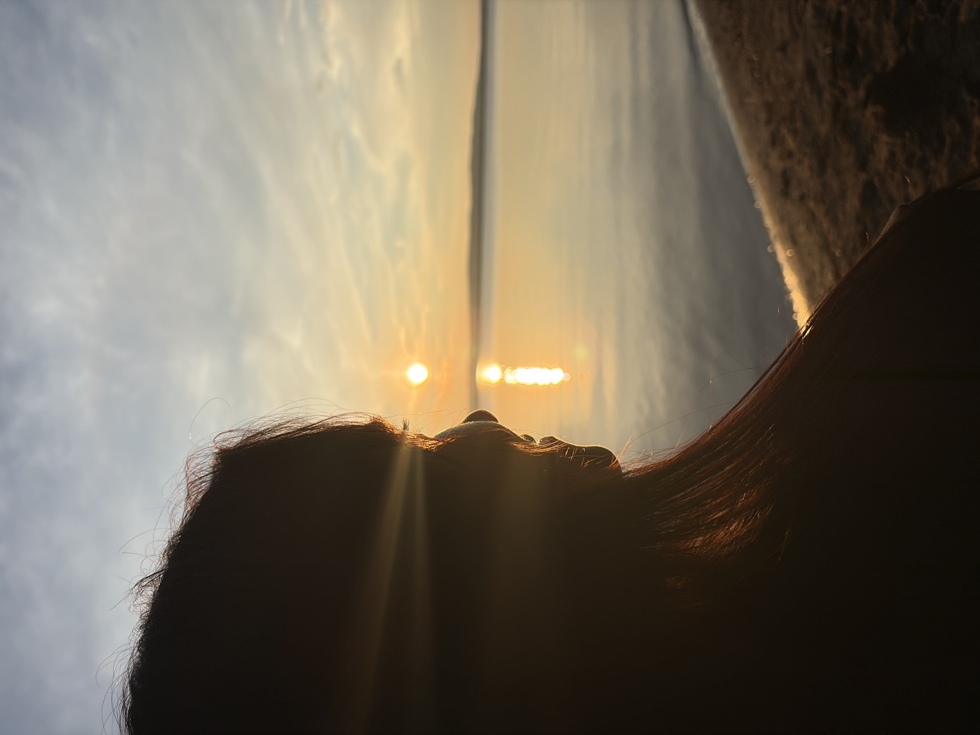 The image shows a silhouette of a person's head and shoulder from the side, facing a body of water during sunset. The sun is low in the sky, casting a warm glow on the scene and reflecting on the water's surface. The person's hair is dark and appears to be long, with a few strands catching the light. The background features a calm body of water, likely a lake or a wide river, with a faint outline of land in the distance. A sandy shore is visible in the foreground, separating the water from the person. The sky is a gradient of soft blues and yellows, with wispy clouds. The overall atmosphere is serene and contemplative. The image was taken in Chaikovsky, Russia, known for its picturesque landscapes and proximity to the Kama River.