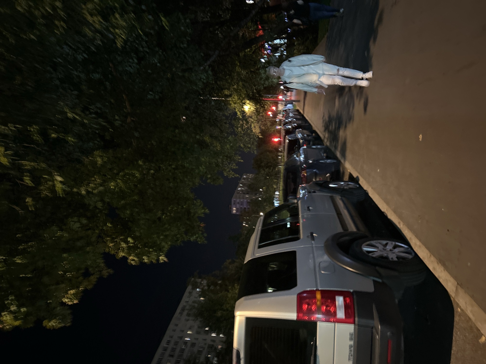 An outdoor, nighttime shot captures a street scene in Moscow, Russia. In the foreground, the rear of a silver SUV is visible on the left. The street is lined with parked cars on the left, and a sidewalk on the right. A person walks along the sidewalk, dressed in light-colored clothing. Several trees with lush foliage overhang the sidewalk and street, casting shadows. In the background, apartment buildings with illuminated windows are visible, along with streetlights and traffic signals. The overall impression is a quiet urban night.