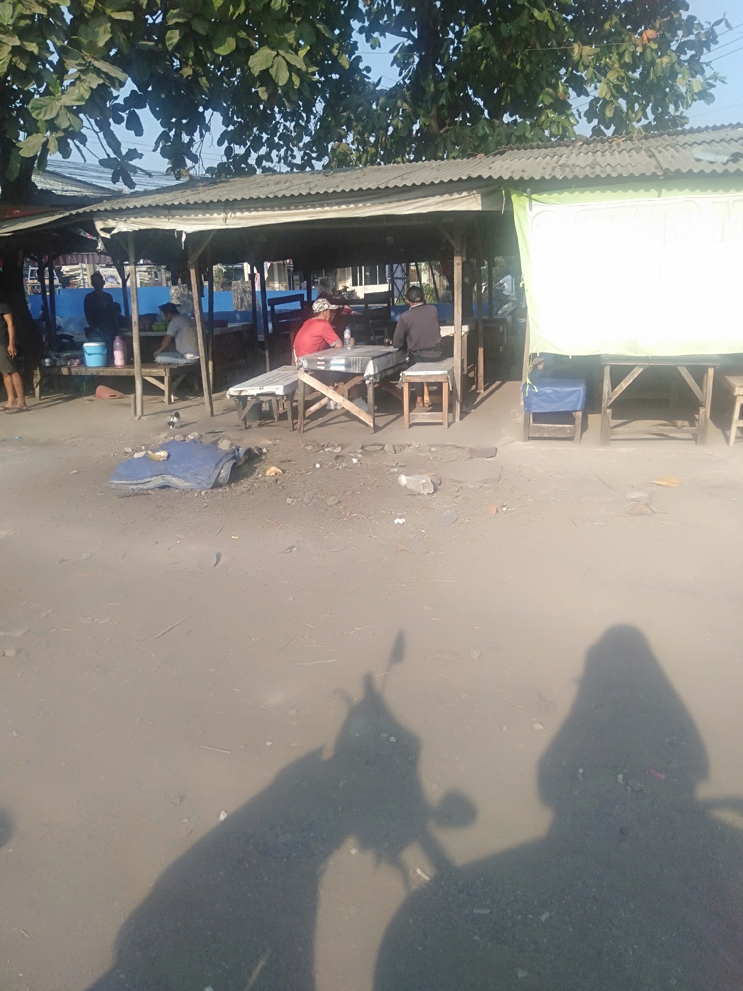 The image depicts an outdoor area with a corrugated metal roofed structure, likely a roadside stall or eatery, located in Sukadami, Indonesia. Sunlight illuminates the scene, casting long shadows. Several individuals are present, with three men seated at tables under the shelter. One man in a red shirt sits at a table on the right, facing left. Another man in a dark shirt is seated on a stool to the left of the first man. A third person, partially visible, is seated at a table in the background to the left. In front of the structure, a blue tarp lies folded on the ground, with some debris and small objects scattered around. A small bird stands on the tarp. The ground is uneven and appears to be dirt or gravel. The shadows of a person or people and a motorcycle are cast across the foreground. The overall impression is one of a casual, possibly open-air dining or resting spot.