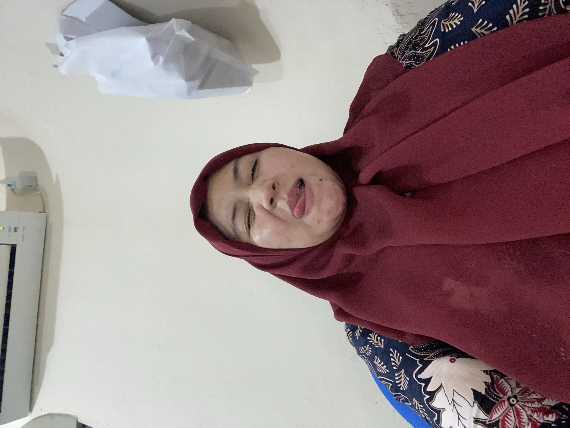 In an indoor setting, a person wearing a maroon hijab and a dark blue shirt with a floral pattern is visible from the chest up. The person's eyes are closed, and their tongue is sticking out in a playful manner. The background consists of a light-colored wall with an air conditioning unit mounted at the top. A white, crumpled object hangs on the wall to the right of the person. The scene appears to be located in Banjarbaru, Indonesia.