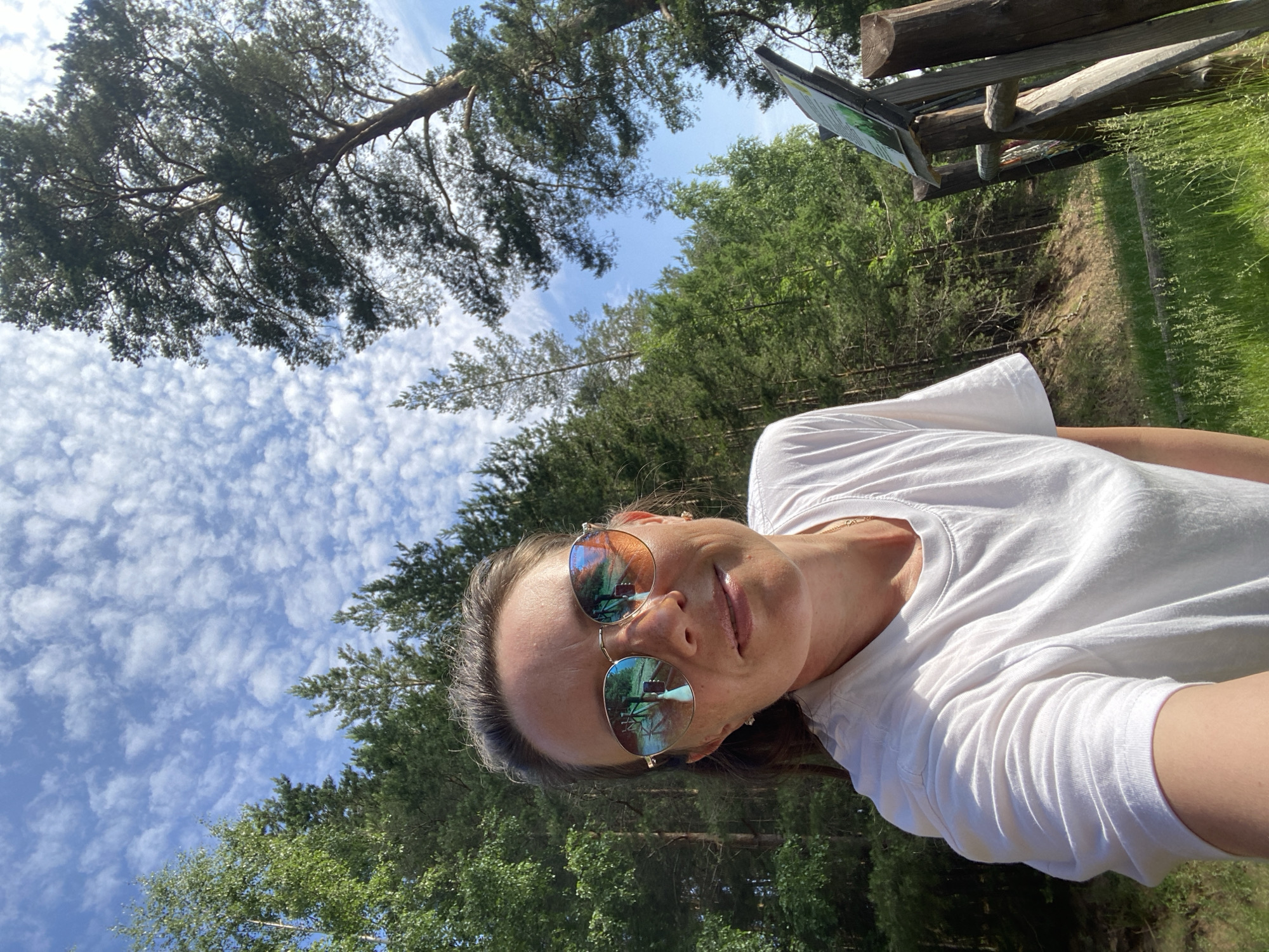A woman is taking a selfie outdoors, with a forest and a cloudy sky in the background. She is wearing a white t-shirt and sunglasses, and her hair is pulled back. There is a wooden fence and a signpost to her right. The scene appears to be a hiking trail or park. The location is the city of Chaykovsky, Russia.