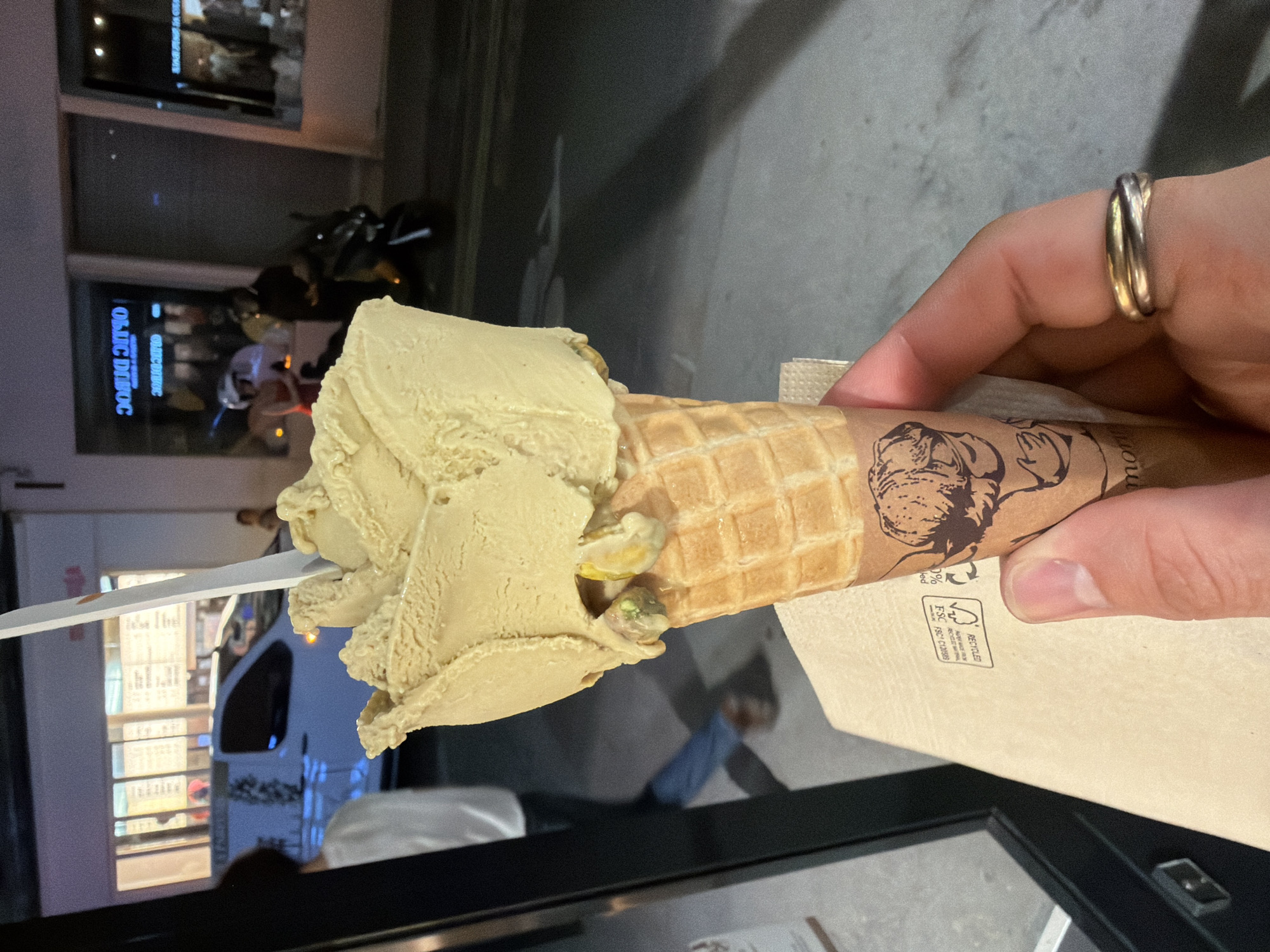 A hand, adorned with a three-band ring, holds a pistachio ice cream cone in front of a Parisian backdrop at dusk. The cone, featuring a waffle pattern, is wrapped in a paper sleeve with a printed logo. The ice cream is melting. A spoon rests on the left side of the cone. The background reveals a Parisian street scene, including a storefront with reflective windows, a scooter, and a white car. The location is Paris, France.