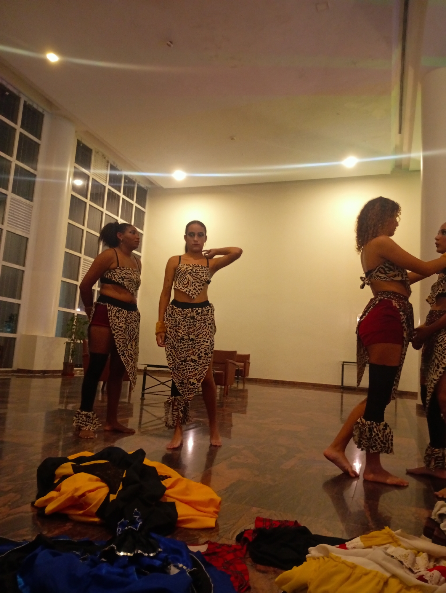 The scene depicts four women in similar costumes, appearing to be practicing or preparing for a performance. They are barefoot and clad in animal print tops and skirts or shorts with matching ruffled anklets. The costumes suggest a tribal or exotic theme. The women are standing in a large, well-lit room with a polished marble floor that reflects the overhead lighting. One woman has her hand on the shoulder of another, potentially adjusting her costume. In the foreground, there is a pile of assorted clothing in yellow, black, and blue colors. The environment appears to be an indoor space, possibly a hotel lobby or rehearsal area, with large windows visible on the left side. The location is in the city of Pedro González, Venezuela. The focus is on the women and their costumes, indicating a performance or artistic preparation in progress.