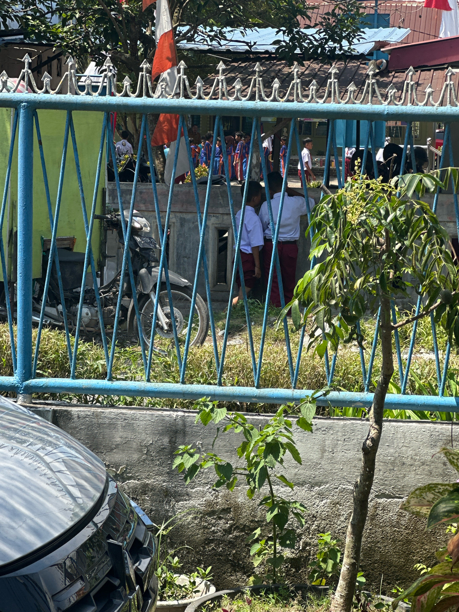 The image captures a view through a blue metal fence at what appears to be a school or community gathering in the City of Binjai, Indonesia. In the background, several children in white shirts and red or blue bottoms are visible. Some children are gathered in small groups, while others are walking. A black motorcycle is parked behind the fence. The environment includes green foliage, a concrete wall, and buildings with tiled roofs. The scene suggests an outdoor event or daily school activities.