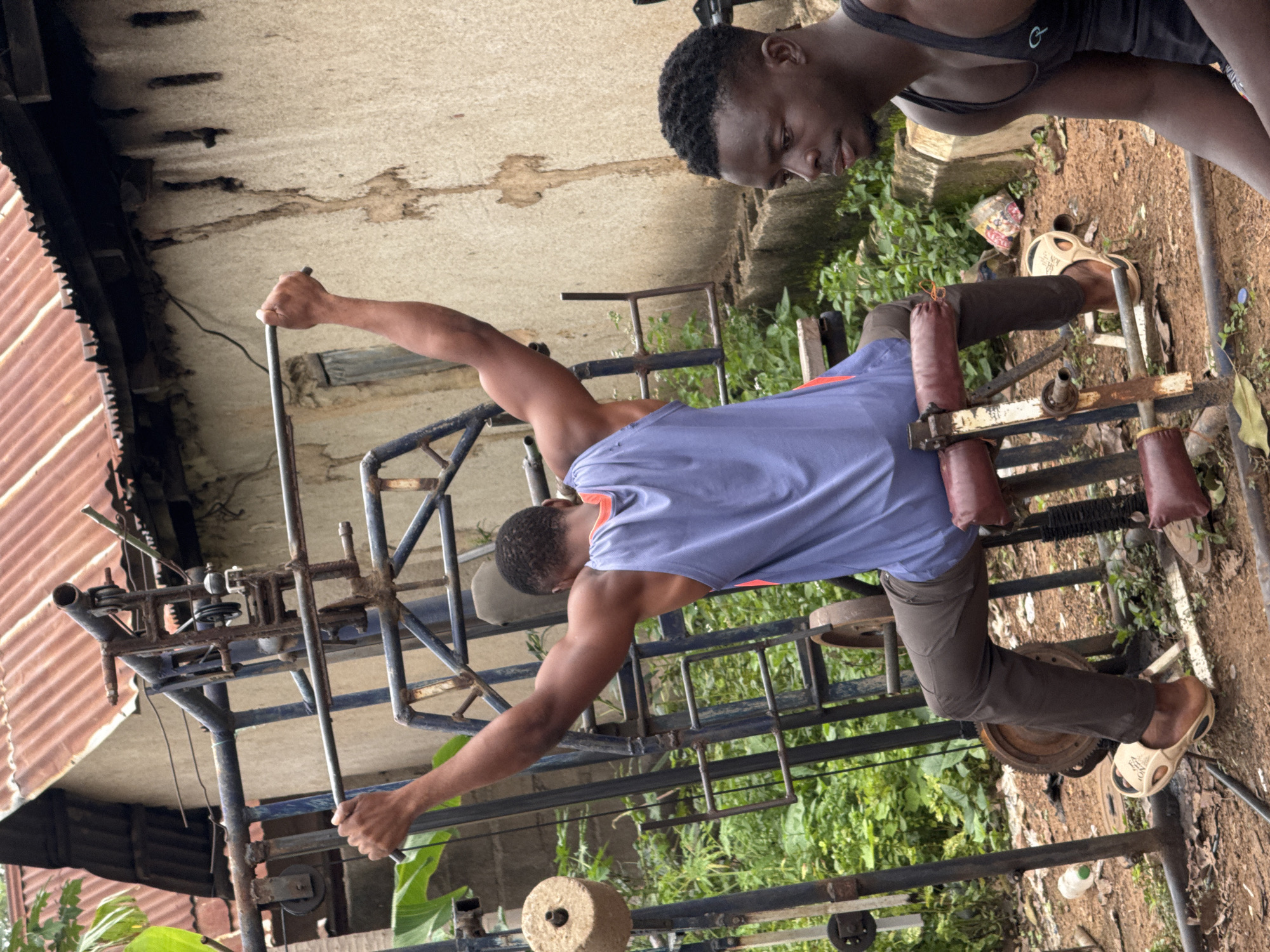 In Akwanga, Nigeria, an outdoor gym setup features two young men. One man is seated on a weight machine, performing a lat pulldown exercise with his arms extended overhead. He is wearing a blue tank top and brown pants. The other man is sitting to the right, watching. He is wearing a black tank top. The gym equipment appears homemade and rusty, situated in a dusty, outdoor area with overgrown vegetation and a corrugated metal roof structure in the background.