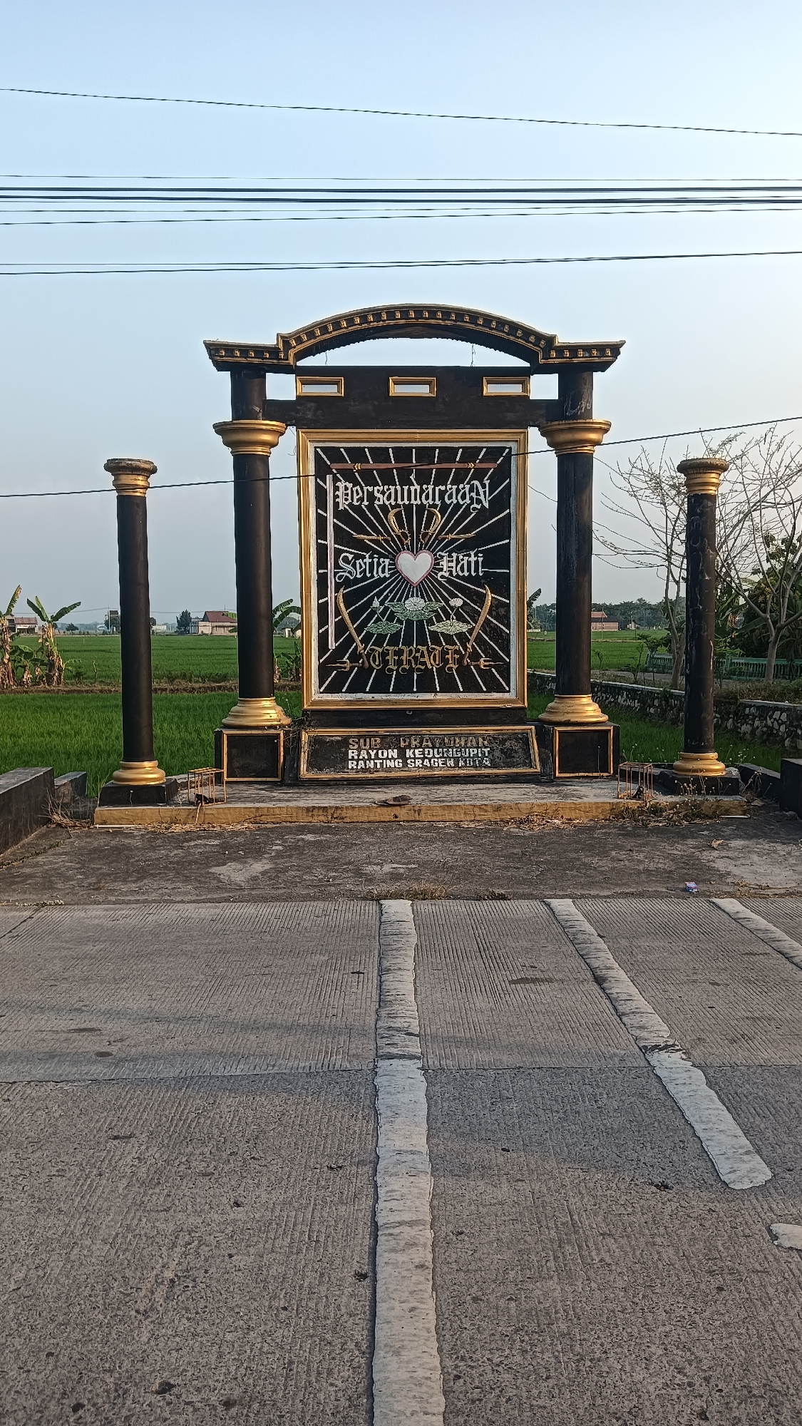 The image shows a monument with the words "Persaudaraan Setia Hati Terate" written on it. The monument is located in Kedungupit, Indonesia, in front of a rice field. The monument is black and gold and has two pillars on either side. In front of the monument, there is a road with white lines. The sky is clear and blue. The overall scene is peaceful and serene.