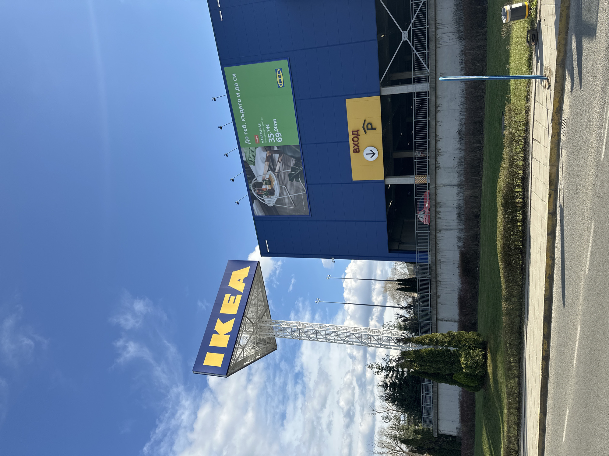 The image depicts an IKEA store, presumably located in Sofia, Bulgaria, based on the presence of Cyrillic text on the building. The view is from the street level, showing the store's exterior. A large blue IKEA building is visible, adorned with a green advertising banner and a yellow parking entrance sign (ВХОД "Entrance," with a parking symbol). Prominently featured is the IKEA logo on a tall white skeletal tower structure. The sky above is blue with scattered clouds. In the foreground, there is greenery with bushes and trees separating the building from the road. A grey pavement road runs along the bottom of the image.