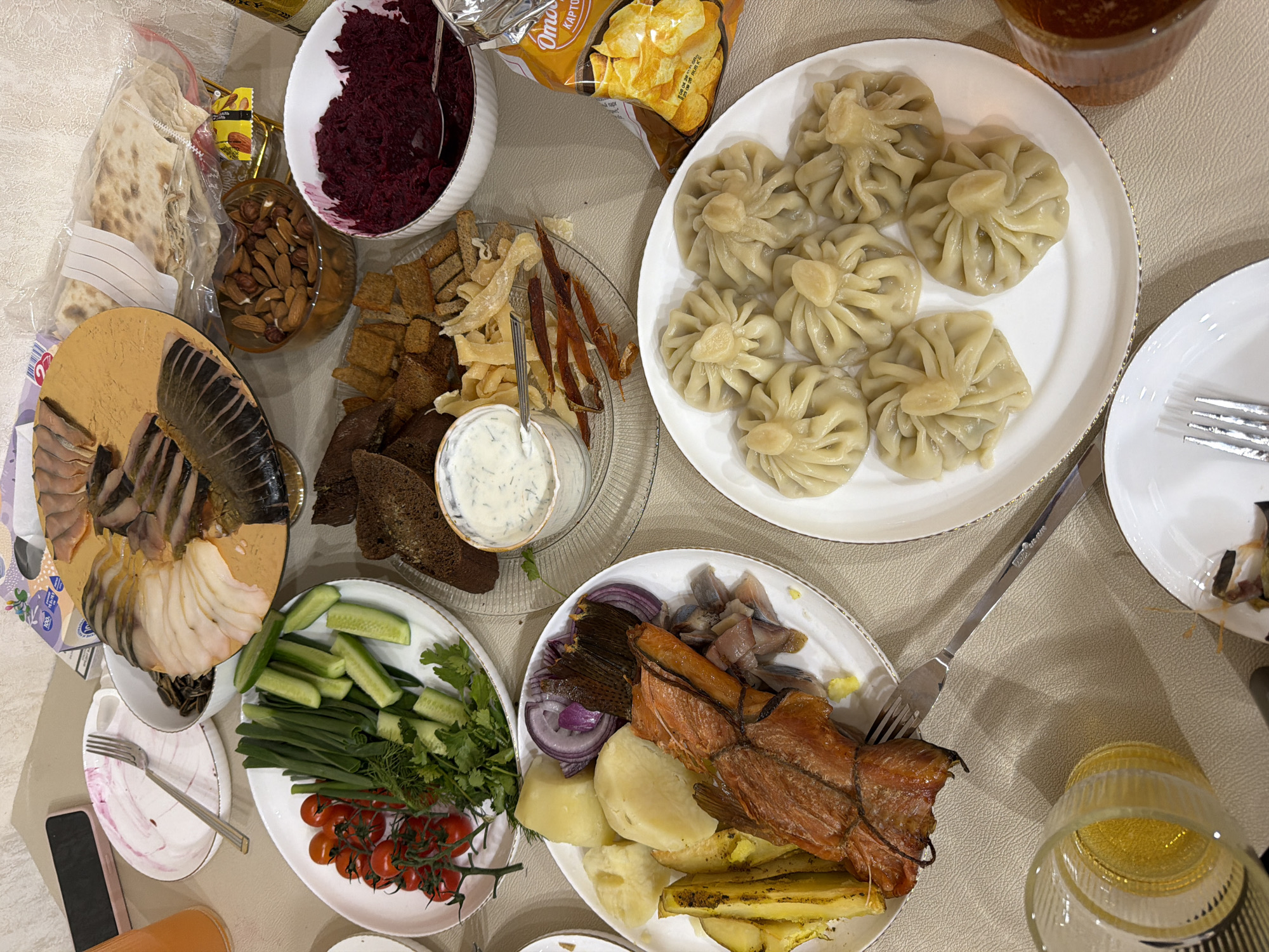 The image shows a table laden with food. There's a plate of what appear to be dumplings, a plate of various types of smoked fish, a plate with vegetables (cucumbers, cherry tomatoes, green onions, herbs), a plate of potatoes, red onion, and more smoked fish. There's also a plate with rye bread and dried fish with a dip. There is a bowl of beet salad, a bowl of almonds, flatbread in a plastic bag, and a bag of chips. The scene suggests a meal or gathering. There is no information in the image to identify this location as Chaikovsky, Russia.