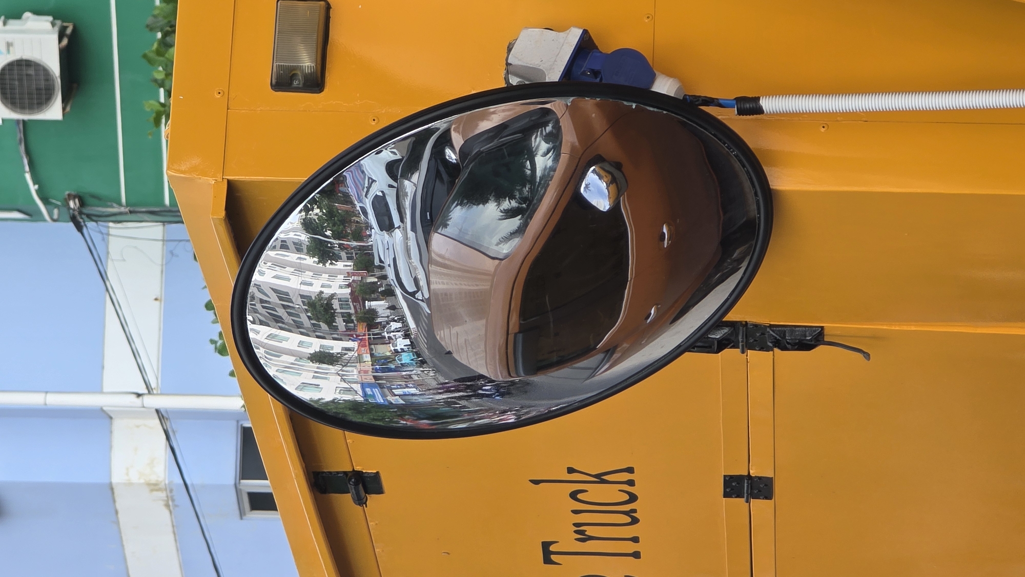 A convex mirror attached to the yellow side of a truck reflects a street scene in Bac Nha Trang Ward, Vietnam. Buildings, vehicles, and other structures are visible in the mirror's reflection. An air conditioning unit is fixed to a green building in the upper left. A white corrugated pipe runs horizontally across the upper right of the truck's side. Black lettering on the truck reads "Truck".