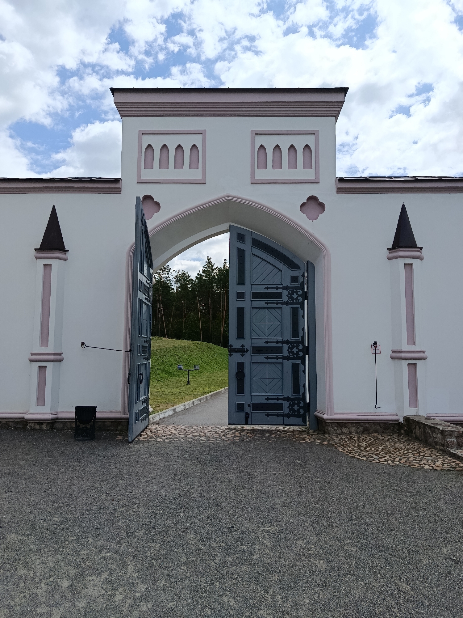 A wide-angle, eye-level shot captures an open, ornate, grey, metal gate at the entrance to a historical site in Kvasavichy Sel'ski Savet, Belarus. The gate is set within a white, stucco wall with pink decorative trim. The wall features gothic-style arched windows and triangular turrets topped with dark conical roofs. The gate, which is partially open, reveals a well-maintained grassy area with a pathway and a dense pine forest in the background. The ground in front of the gate is covered in dark grey gravel, transitioning to cobblestones near the entrance. The sky above is a mix of blue with scattered white clouds. There are no people or animals visible in the scene.