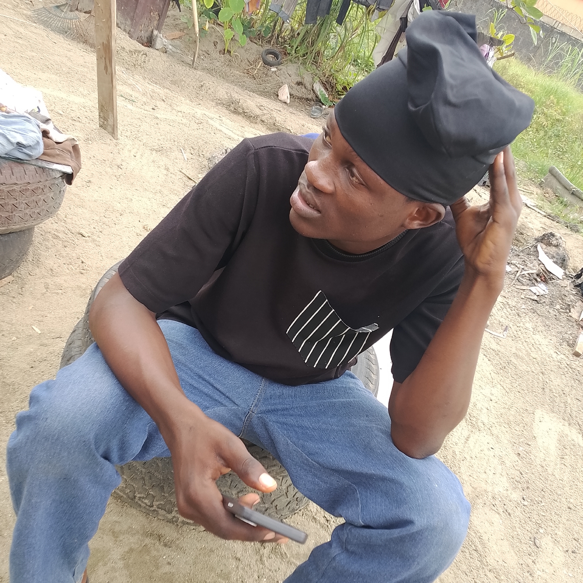 A young Black male is pictured from the chest up, wearing a black t-shirt with a white-striped pocket, blue jeans, and a black do-rag. He is seated outdoors on what appears to be a stack of tires. His right hand is placed on his head, and his left hand is holding a cell phone. The background is sandy with some vegetation and what looks like drying clothes hanging on a line. The scene is likely in Ado, Nigeria, given the environmental context and the appearance of the individual.