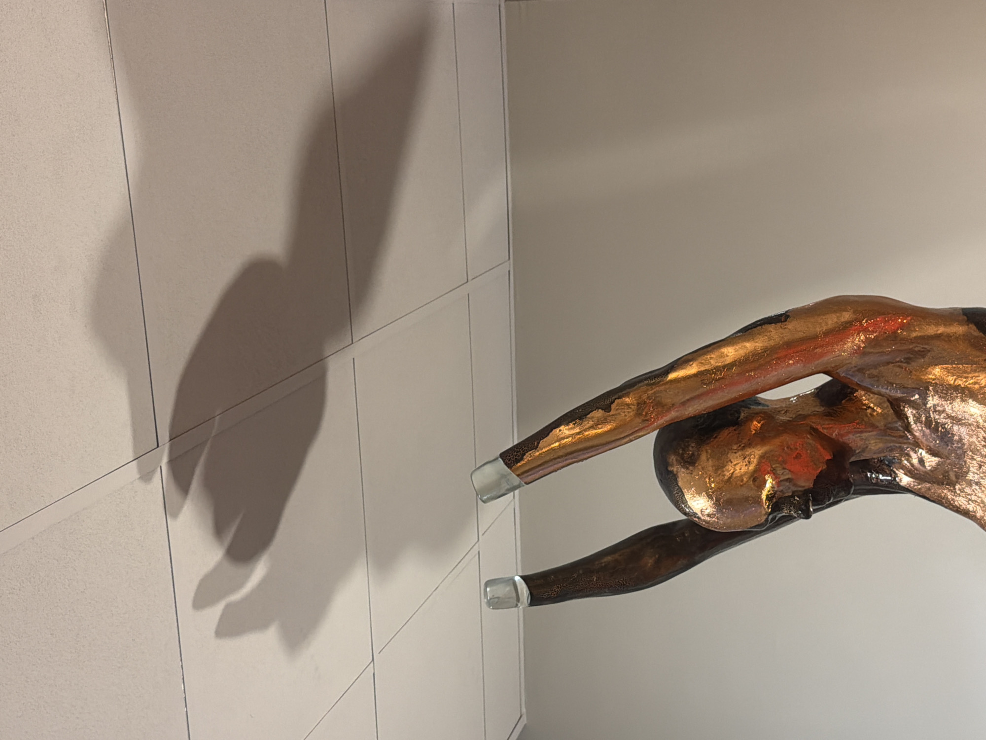 A modern sculpture of a human figure, predominantly golden and bronze-toned, is displayed against a neutral background. The figure is depicted in a dynamic pose, appearing to be mid-dive, mid-leap, or mid-swim, with arms extended forward and the body elegantly arched. The metallic surface of the sculpture exhibits a textured, almost flaky or hammered finish, showcasing a luminous blend of copper, gold, and darker bronze hues, indicating a rich patination. Notably, the extremities of the figure's limbs, specifically the hands and possibly feet, are crafted from a clear, translucent material, such as glass or acrylic, providing a striking contrast to the opaque metallic body.
The sculpture is situated in an interior setting against two light-colored walls. To the left, a textured wall with visible vertical and horizontal grout lines suggests a tiled or paneled surface. A prominent, elongated shadow of the figure is cast onto this wall, mirroring its energetic form. The wall to the right and behind the sculpture is smooth and unadorned, serving as a minimalist backdrop that allows the artwork to stand out. The lighting is indirect, accentuating the metallic sheen and the sharp definition of the shadow. There is no visible text in the image. This artwork is located in Milan, Italy.
