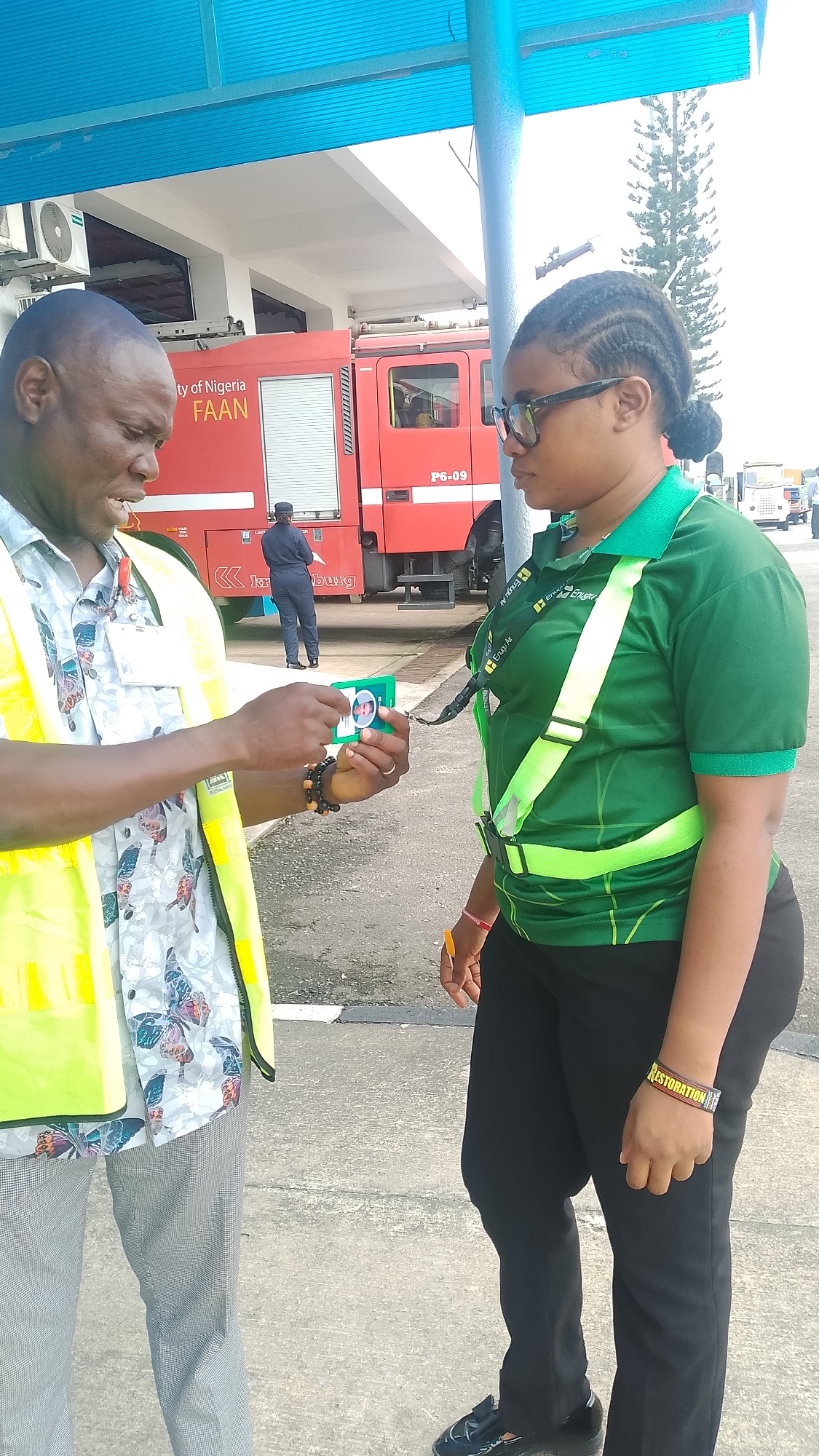 In Orerokpe, Nigeria, a man wearing a yellow high-visibility vest over a patterned shirt is handing a small green card with a picture on it to a woman. The woman is wearing a green collared shirt with a high-visibility harness over it and black pants. Behind them, a red fire truck is parked, with "ty of Nigeria FAAN" visible on its side. The scene appears to be outdoors, possibly at an airport or similar facility, with a paved ground and a building with a blue overhang visible.