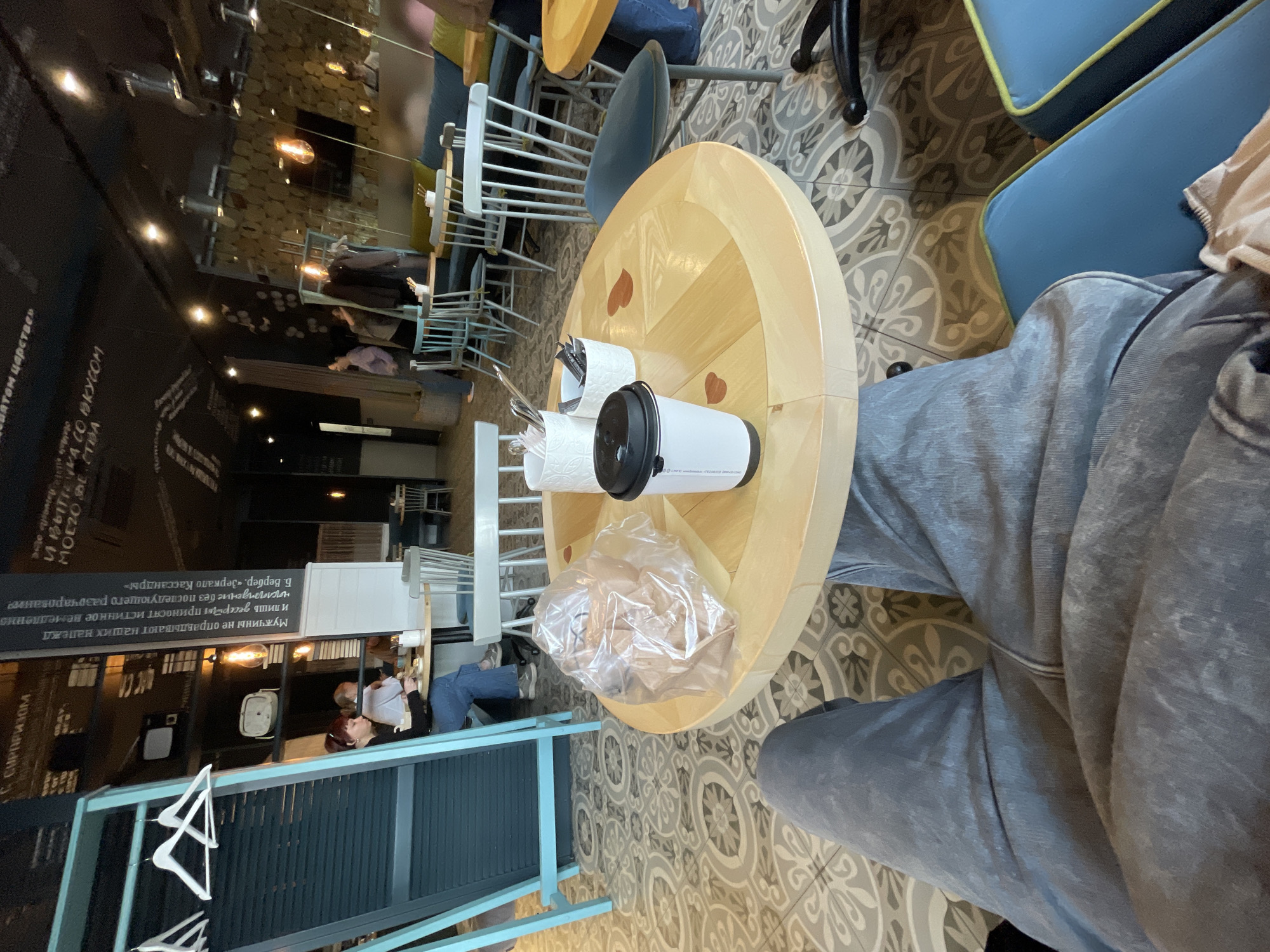 Here is a detailed description of the image:
**Scene:** An indoor view, likely a café or restaurant, centered on a round wooden table with objects on it. The perspective is from an elevated point of view, almost directly above the table.
**Location:** This is probably in Krasnoyarsk, Russia.
**Objects:**
* A round wooden table with heart-shaped decorations.
* A white to-go coffee cup with a black lid on the table.
* Plastic cutlery and condiments packaged for take-away.
* A partially visible blue chair next to the table.
**People:**
* One person, wearing grey sweatpants, is sitting at the table. This is only partially visible from the lap down.
**Actions/Interactions:**
* The person is seated at the table, possibly about to enjoy a meal or drink from the items on the table.
**Environment:**
* The café has a patterned tile floor.
* Other tables and chairs are visible in the background.
* The walls have decorative elements, including what looks like a chalkboard with writing in Russian.
* Hanging clothes from the ceiling.
* Brightly lit by natural light.