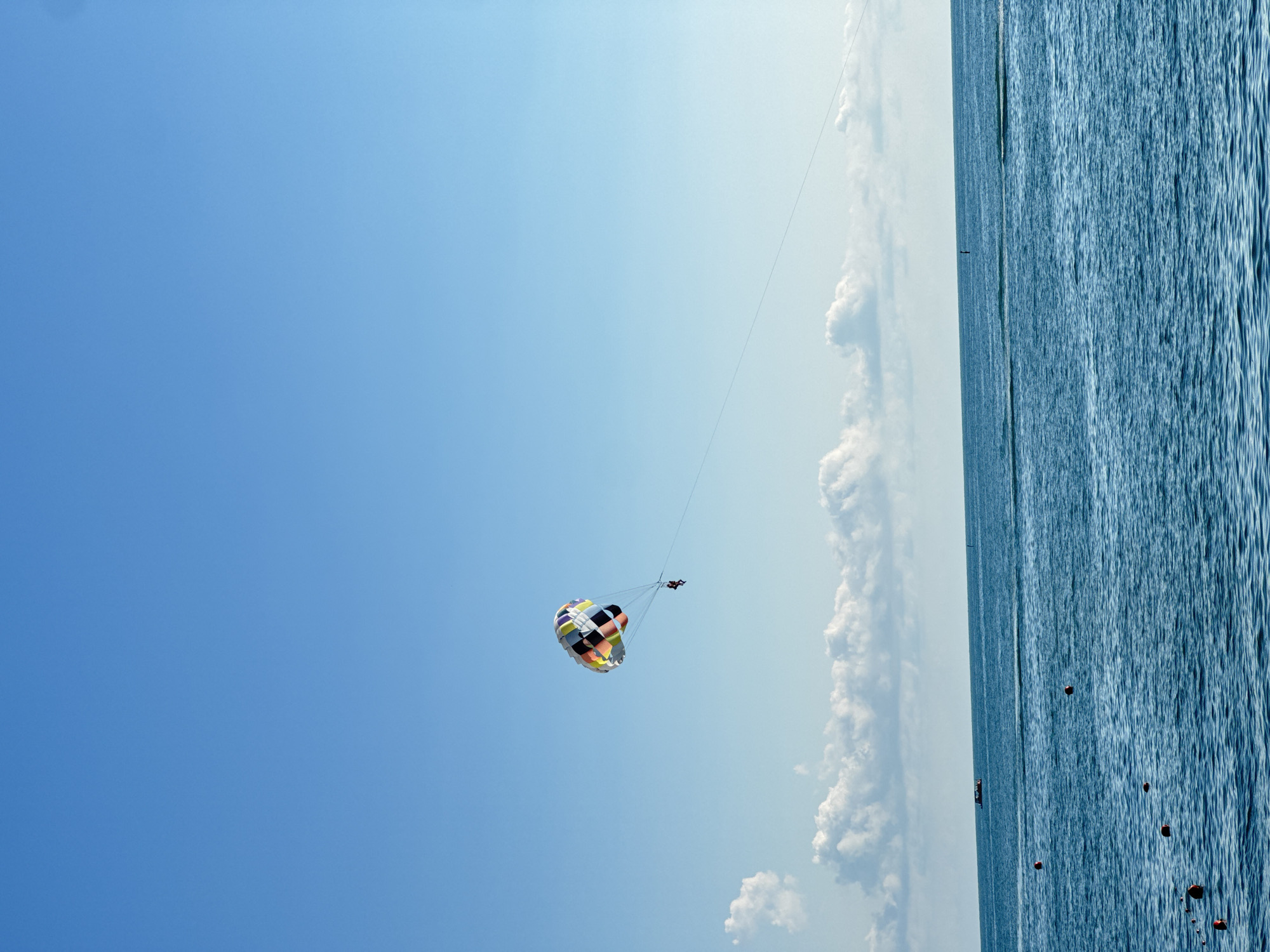 This media file shows a parasailer in the sky above the sea. The parasail is colorful, with a checkered pattern of blue, yellow, red, green, and white. Two people are visible, one attached to the parasail and another person in a boat on the water below. The water is a deep blue and appears to be choppy. The sky is clear and blue, with a few white clouds. The sun appears to be shining, suggesting it is daytime. The location is Sochi, Russia. The activity is parasailing, a recreational activity where a person is towed behind a boat while attached to a parachute-like canopy. The overall scene evokes a sense of leisure and adventure.