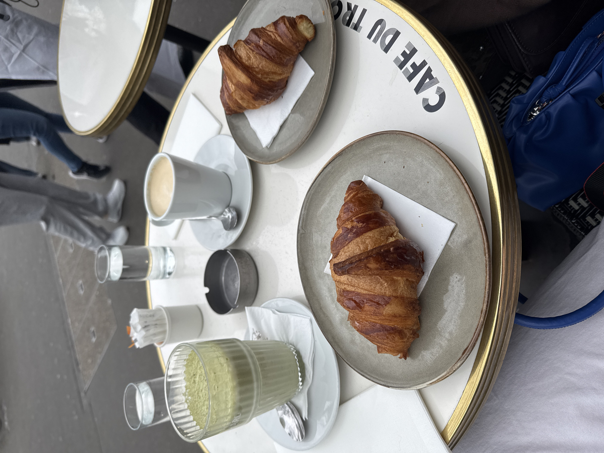 The image depicts a table at the "Café du Trocadéro" in Paris, France, judging by the inscription on the table. On the table are two plates, each with a croissant, a cup of coffee, and a glass of some sort of beverage on a white plate. There are also sugar packets and a small ash tray. Parts of people and their belongings are visible around the table. The overall scene suggests a casual dining or coffee break setting.