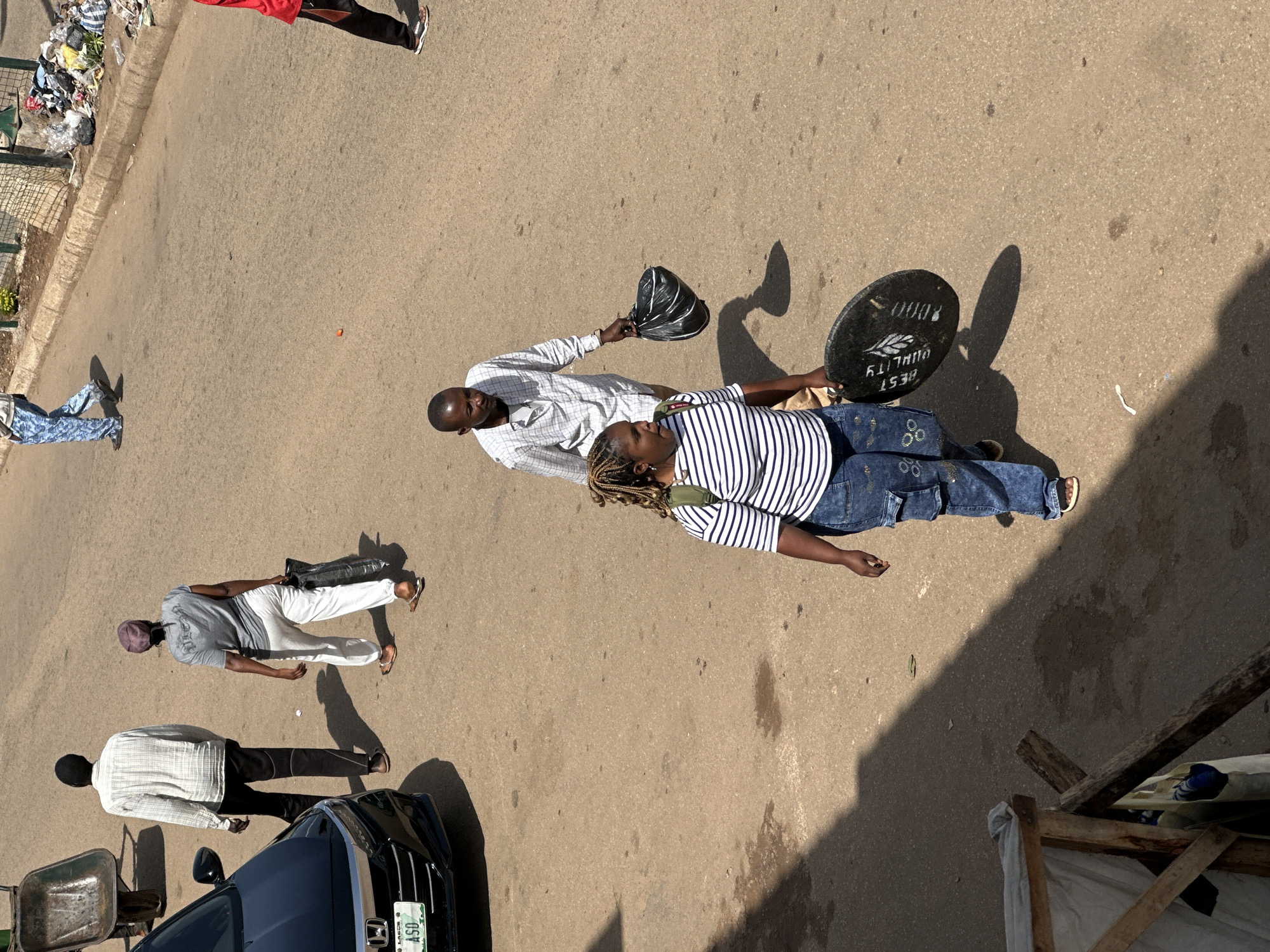In Bukuru, Nigeria, on a sunlit, dusty asphalt road, a group of individuals are captured in motion. A black sedan, possibly a Honda, is parked on the left side of the frame, its license plate bearing "LASG A50". To its right, a man in a checkered long-sleeved shirt and black trousers walks away from the camera. Further down the road, a woman in a gray t-shirt and white pants carries a black bag.
In the foreground, a man in a white checkered shirt and light-colored trousers walks towards the viewer, holding a black bag. Beside him, a woman with braided hair, wearing a striped t-shirt and denim jeans, also walks towards the viewer, carrying a black circular object with white markings that appear to read "BEST QUALITY". A shadowed, makeshift wooden structure is visible in the lower left corner, partially obscured by shadow. In the background, trash is piled near a wire fence, and another person in patterned blue trousers walks away from the camera. The overall scene depicts everyday street activity under a bright sky.