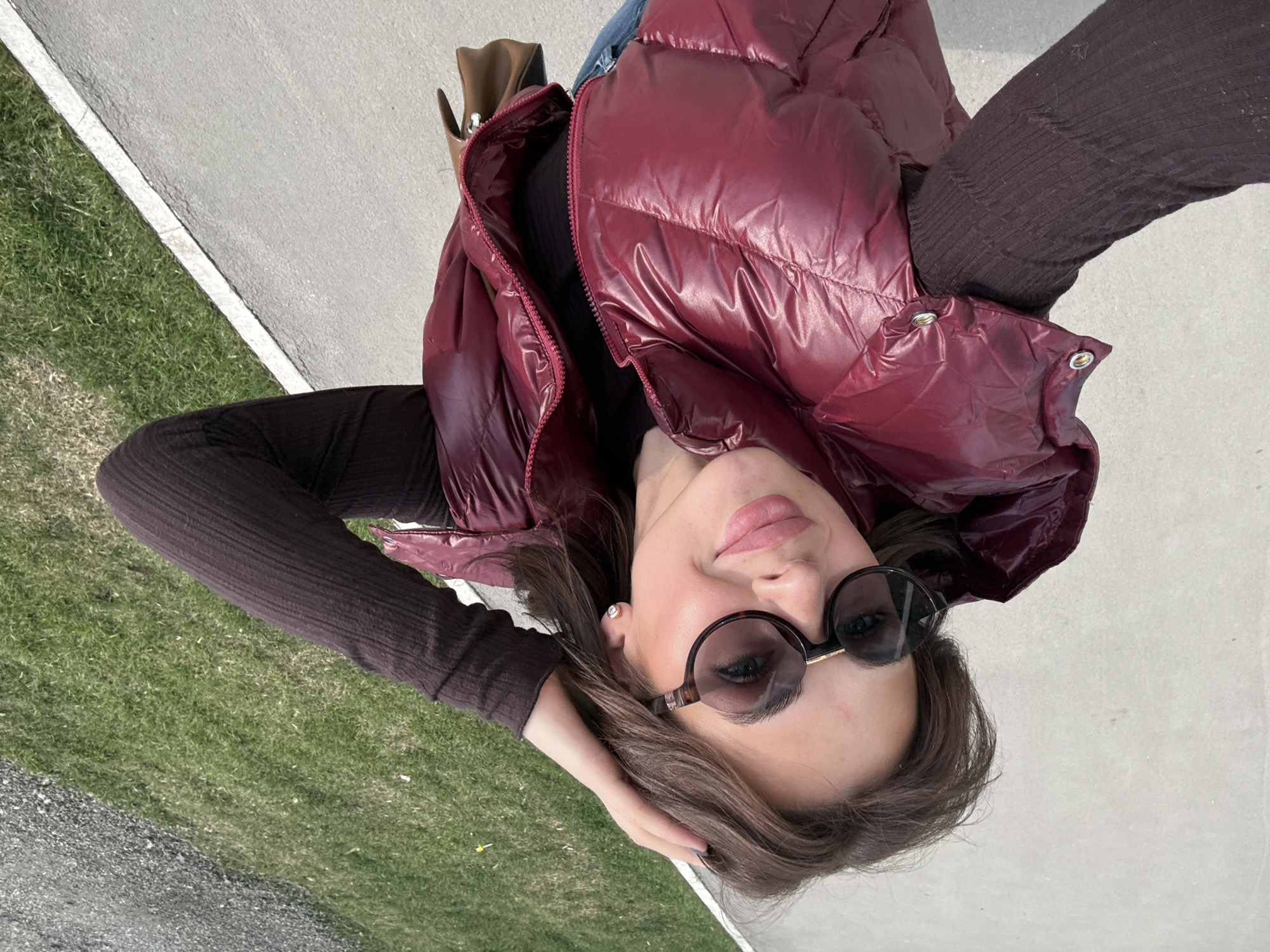 A young woman wearing a maroon puffer vest over a brown long-sleeved shirt and sunglasses is standing on a sidewalk with green grass on the side. The image is taken from a high angle, looking down at her. A brown purse is visible near her shoulder. The location is potentially in Perm, Russia.