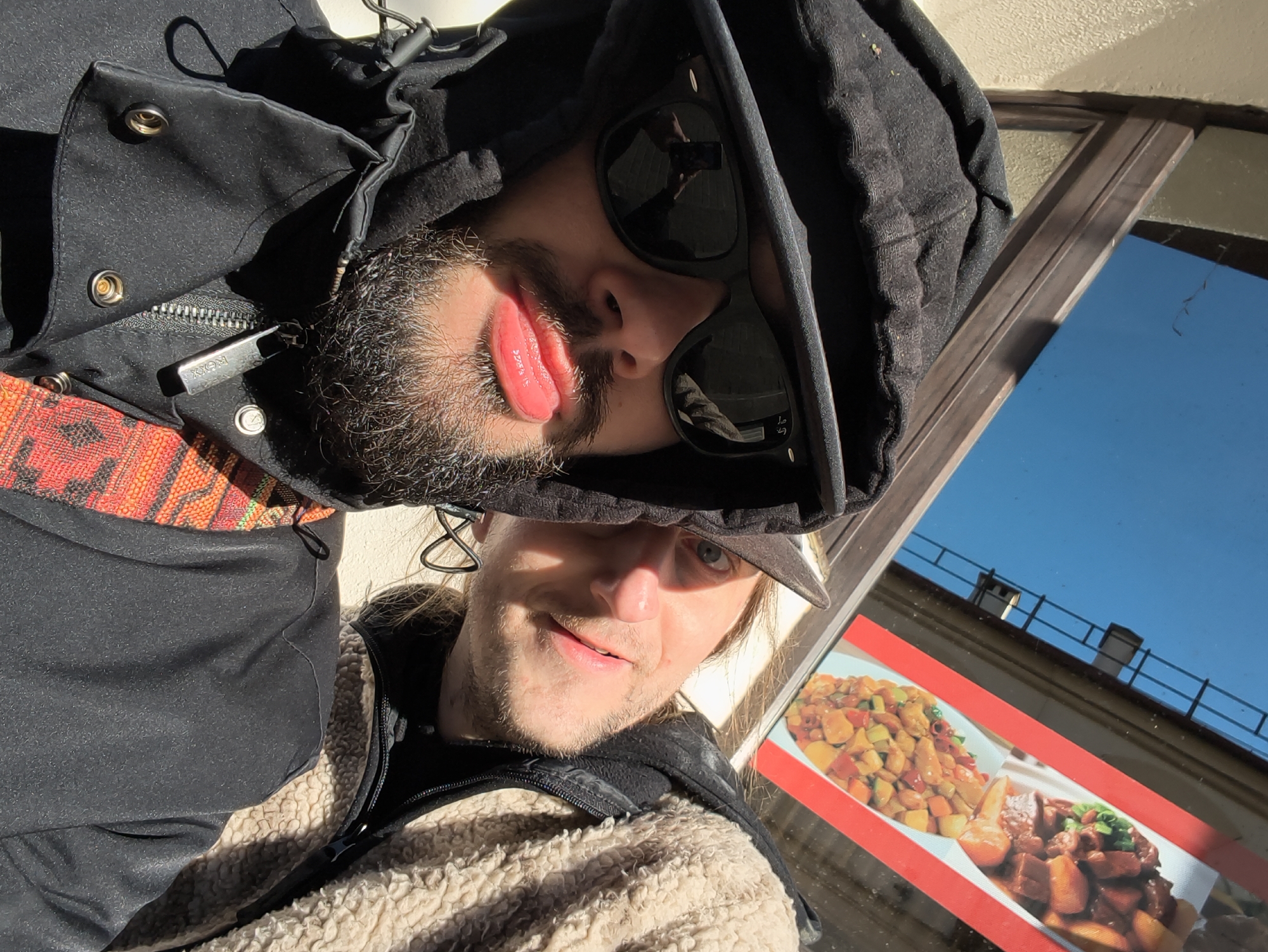 Two men are taking a selfie. One man is wearing a black hooded jacket and sunglasses, with a beard. He is facing the camera sideways and making a kissy face. The other man is wearing a baseball cap and a tan jacket. The reflection of a blue sky and a building is visible in a window behind them. There is a sign for food visible in the window. The location is likely Hrodna, Belarus due to the style of the building in the reflection.