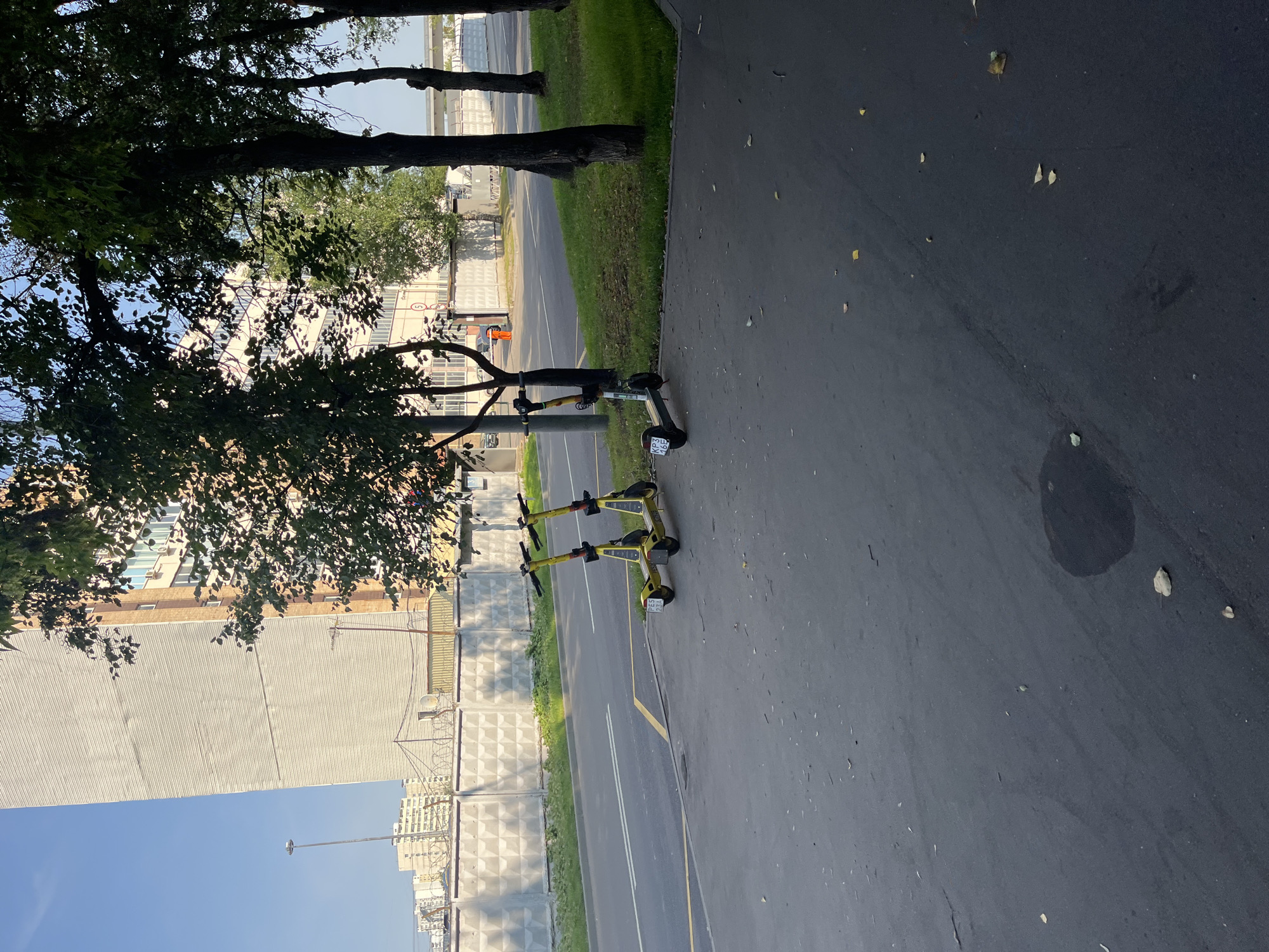 A light gray concrete fence with a diamond pattern runs along the side of a road in Moscow, Russia. Barbed wire is visible on top of the fence. Two yellow electric scooters are parked on the asphalt sidewalk next to the fence. Behind the fence, a tall, light-colored building with many windows stands under a clear blue sky. Trees with green leaves are present in the foreground and between the building and the road. The ground is paved with dark gray asphalt, with some fallen leaves scattered on it.