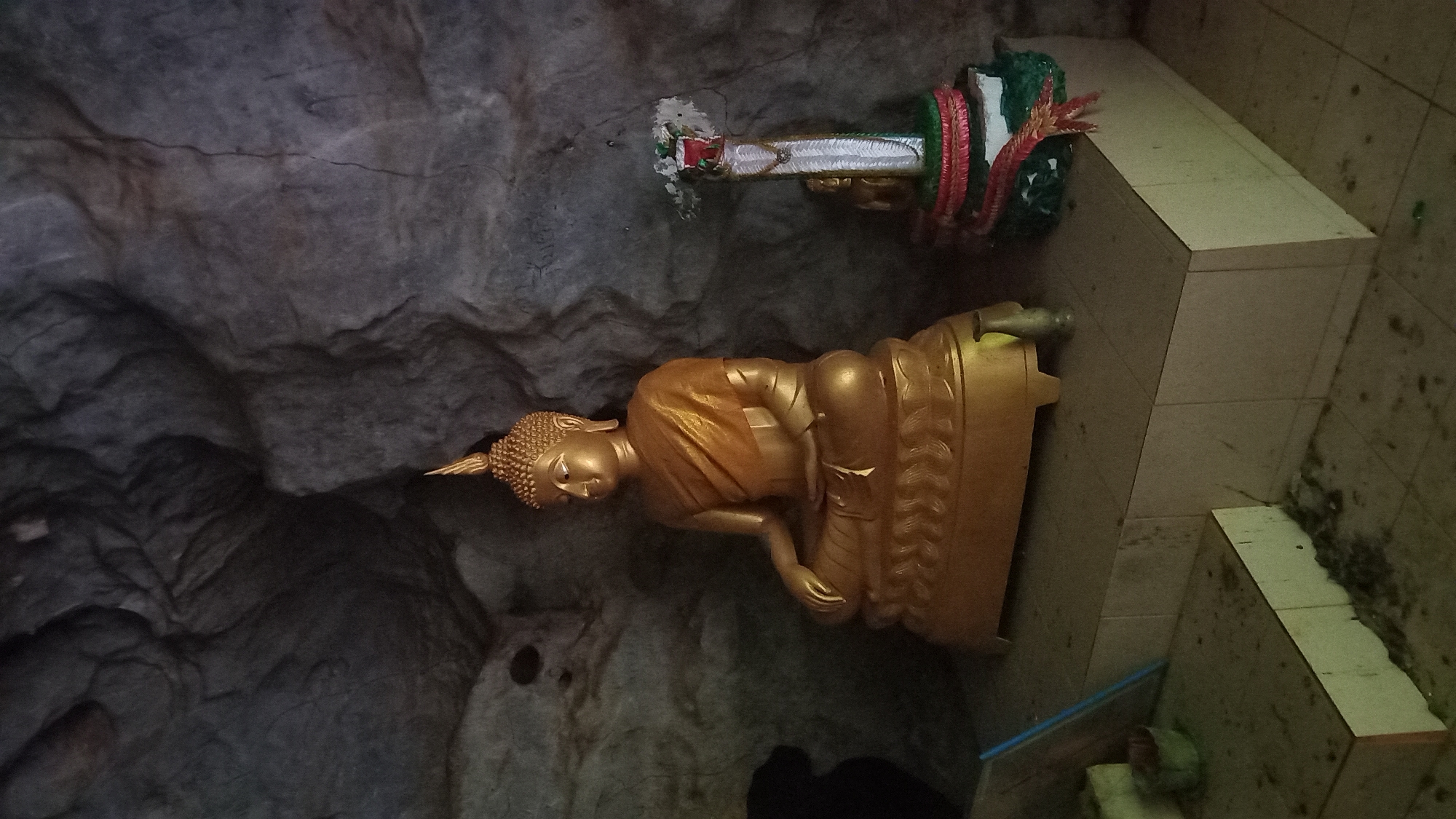 The photograph shows a golden Buddha statue inside a cave. The cave wall is rocky and dark. The Buddha sits on a decorated base, with offerings positioned behind it on a tiled platform. The image was taken at Ban Pak Ra, Thailand.