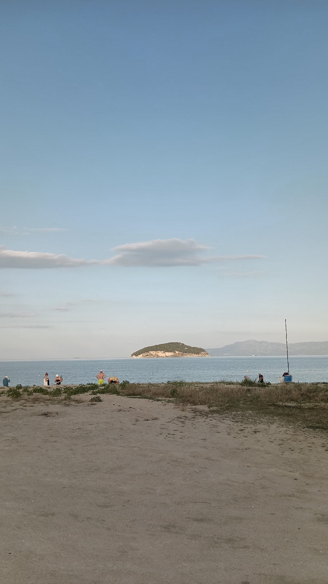 The image captures a serene coastal scene in Nea Irakleitsa, Greece. In the foreground, a sandy beach is sparsely covered with dry grass and some small green plants. Several individuals are scattered along the shoreline. To the left, a person wearing a blue top and dark bottoms is seated. Moving right, three individuals are standing together; two appear to be women wearing light-colored tops and dark bottoms, and one is a person with dark hair wearing a light top and dark bottoms. Further right, a person in yellow shorts stands facing away from the camera, while another person is lying on a yellow object. In the middle ground, the calm, blue sea stretches to the horizon. A small, verdant island with a rocky base is visible in the distance, with mountains forming the backdrop. To the right of the island, a solitary thin pole stands upright in the water, possibly marking a fishing spot or a boundary. In the far right of the frame, two people are near the water's edge, one appearing to be standing and the other possibly seated or bending down. A blue object, possibly a container or chair, is near the standing figure. The sky is a pale blue with a few wispy clouds, suggesting a clear day.