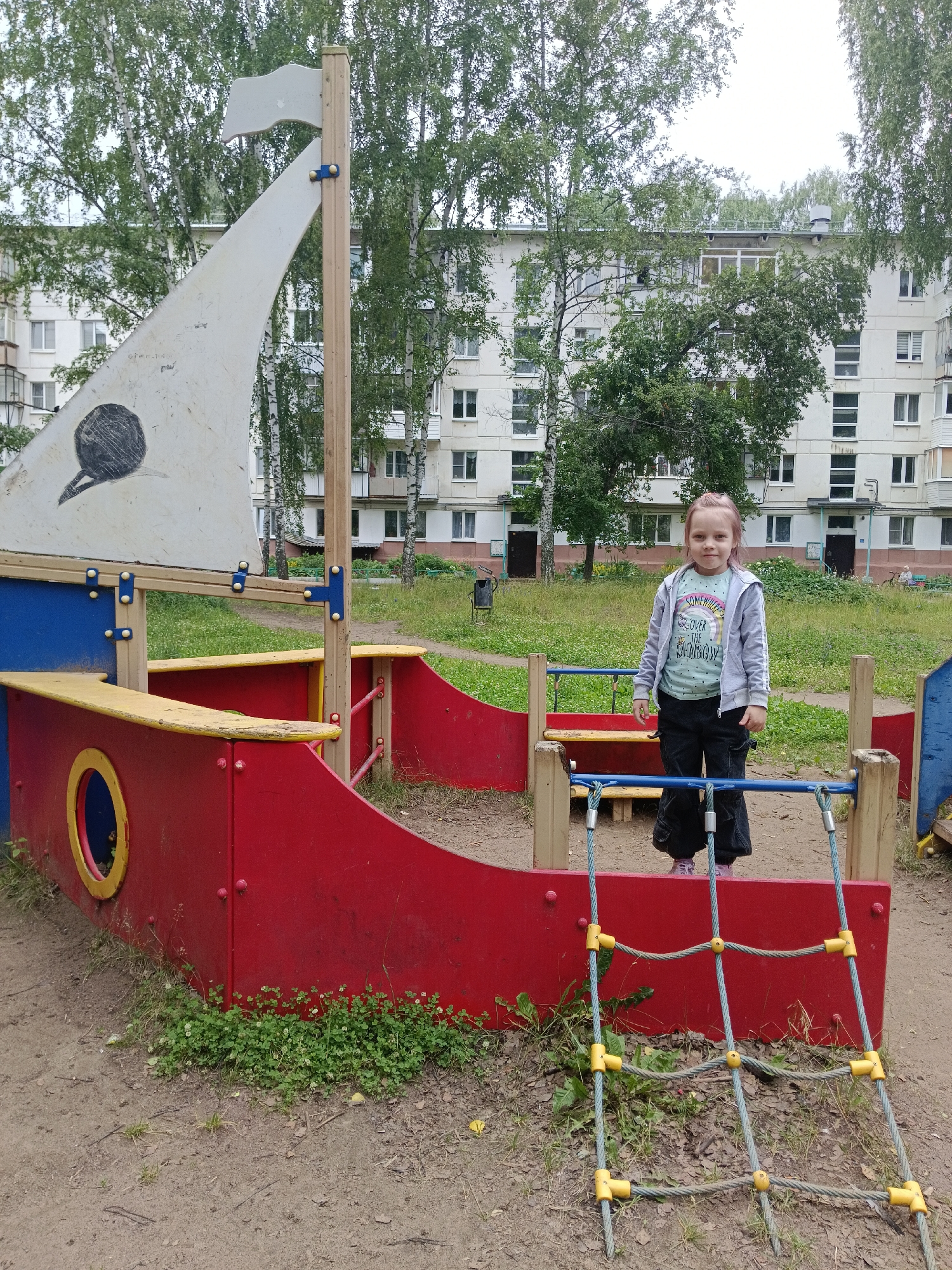 A young girl with pink-streaked hair stands in front of a red and blue pirate ship-themed playground structure. She is wearing a light grey hooded jacket over a mint green t-shirt with text that reads "SOMEWHERE OVER THE RAINBOW," and black pants. The playground equipment is made of wood and brightly colored plastic. Behind the playground, a multi-story, light-colored apartment building is visible, surrounded by green trees. The ground is a mix of dirt and patchy grass. The scene appears to be in a residential area, possibly a courtyard or park. The lighting suggests it is daytime, and the overcast sky implies cloudy or overcast weather. The girl is looking directly at the camera with a neutral expression. The location is identified as Chaikovsky, Russia.