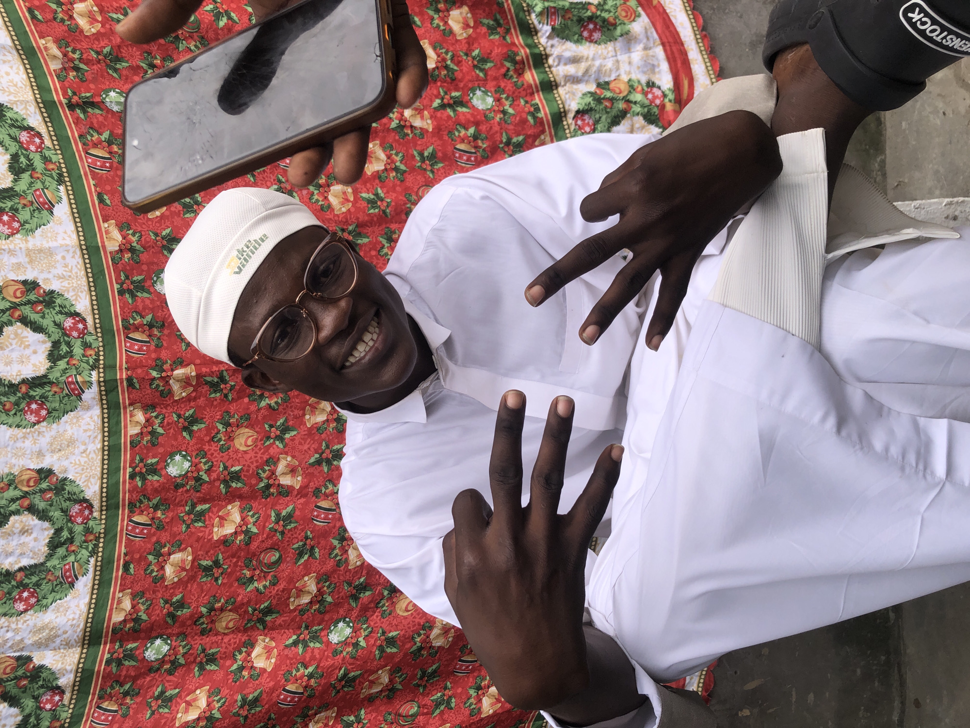 A young man wearing a white thobe and cap with glasses is sitting and posing for a photo. He is smiling and has his hands up, showing four fingers on his left hand and two fingers on his right hand in a peace sign. A phone is held up in front of him by another person's hand, obscuring part of his face. The background is a red cloth with Christmas-themed decorations, including wreaths and bells. The image appears to have been taken outdoors, possibly in Ado, Nigeria, as indicated by the presence of a black flip-flop shoe with "VENSTOCK" written on it, which is a common brand in the region. The lighting suggests it is daytime.