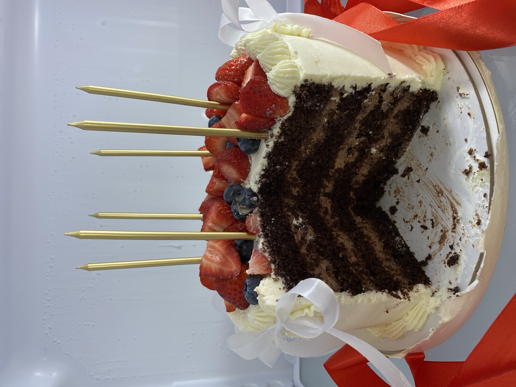 A partially eaten chocolate cake with white frosting and fresh strawberries and blueberries on skewers is in a refrigerator. The cake is decorated with white and red ribbons. The location is not specified and there is no indication of Sochi, Russia.