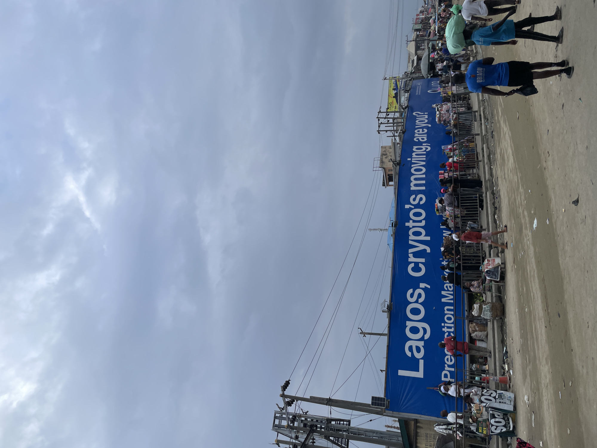 This is a medium-shot, eye-level, outdoor photograph taken on a cloudy day in a commercial area of Lekki, Nigeria. Dominating the scene is a large, bright blue banner with white text that reads "Lagos, crypto's moving, are you?". Below this, in smaller white text, it says "Prediction Market". The banner stretches across the top half of the image.
In front of the banner, a crowd of people are gathered. Some are standing and talking, while others appear to be browsing goods displayed on the ground or in stalls. There is a metal barrier separating the people from the banner. Several individuals are carrying goods, and some are wearing casual clothing. Two men in the foreground are walking away from the camera, one carrying a large green bag on his head.
The ground is dirt and appears somewhat uneven, with scattered debris. Various objects are visible on the ground, including bags, baskets, and what appear to be promotional flyers. Electrical poles and wires are present on the left side of the image, and some buildings are visible in the background. The overall impression is a busy marketplace with a prominent advertisement for cryptocurrency.