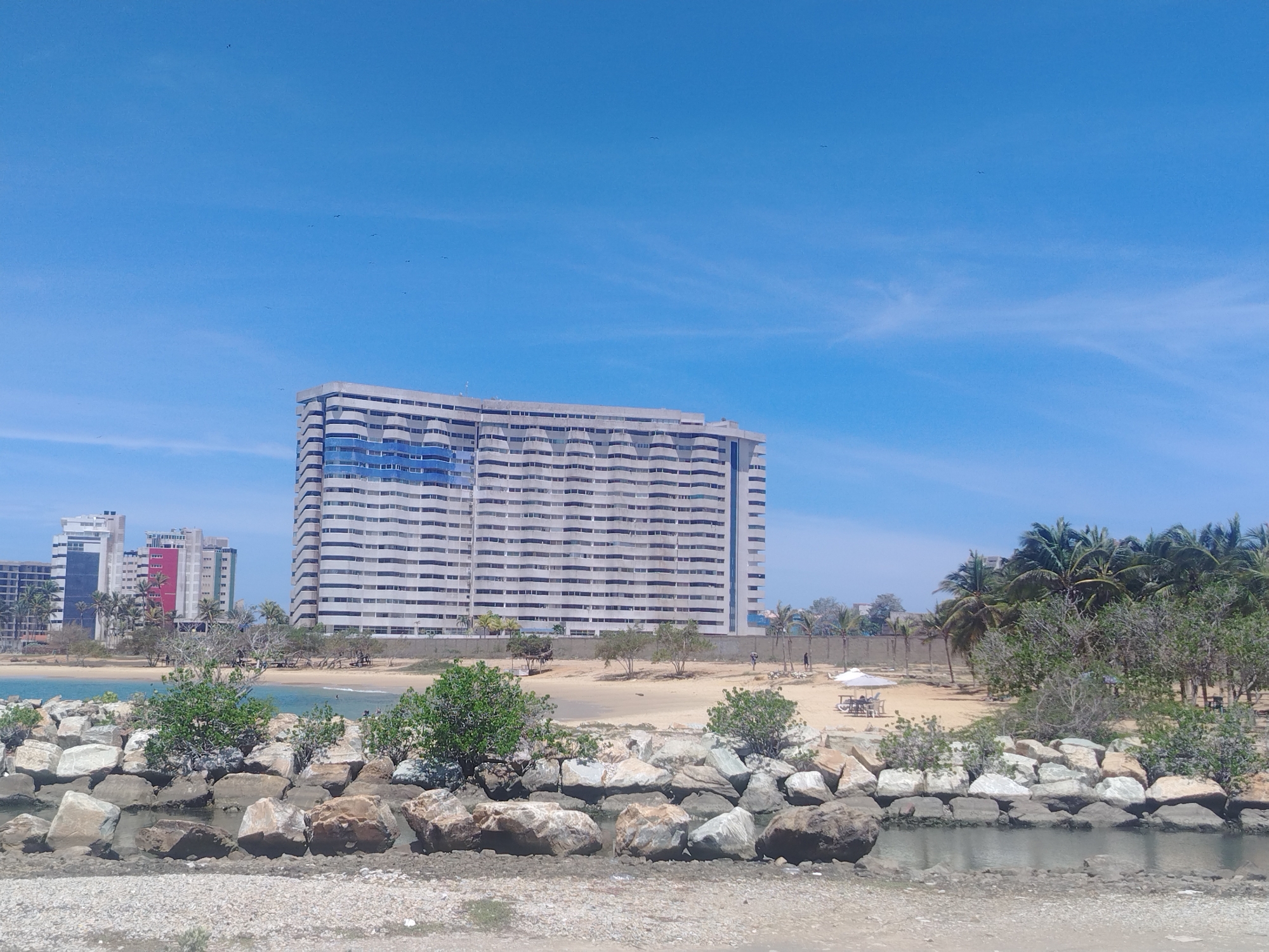 The image shows a coastal view in Porlamar, Venezuela, featuring a large, multi-story white building with blue accents on its facade, situated behind a sandy beach. Several other modern buildings, some with colored panels, are visible in the background. Palm trees and other vegetation are scattered along the shoreline. In the foreground, a rocky breakwater separates the sea from the land, with a body of water visible between the rocks. The sky is clear blue with a few wispy clouds, and several birds are flying in the distance. There are a few people on the beach, one near a white umbrella and picnic table, and others walking. The scene appears to be a sunny day at a tropical urban beach.